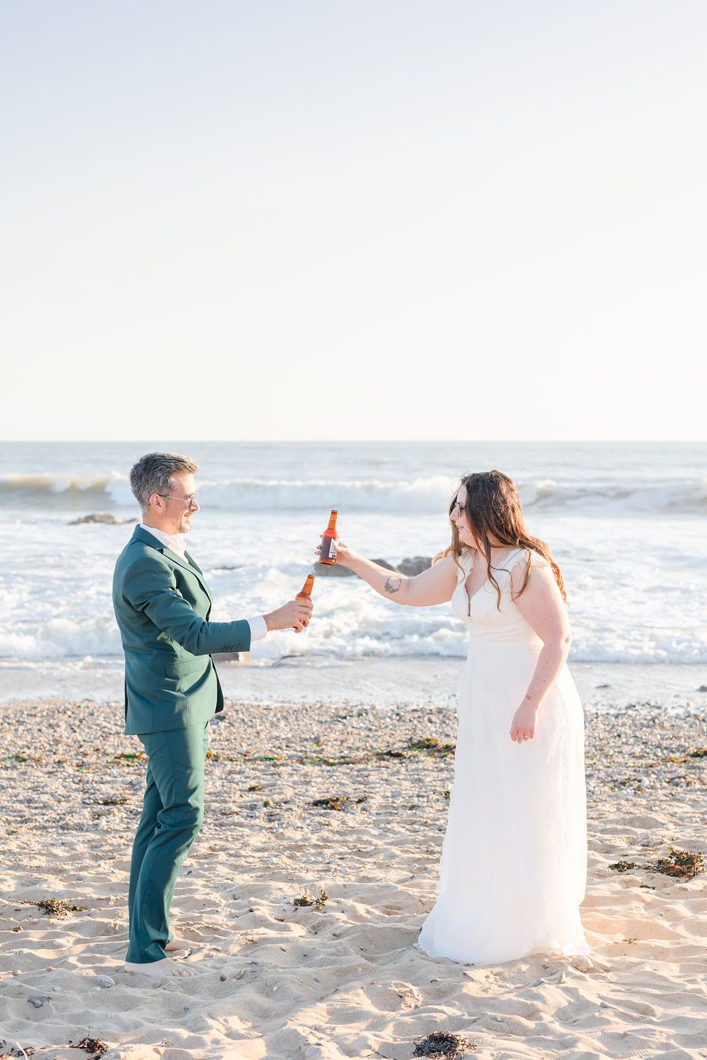 Couple celebrating with champagne on sunny beach during wedding photoshoot.