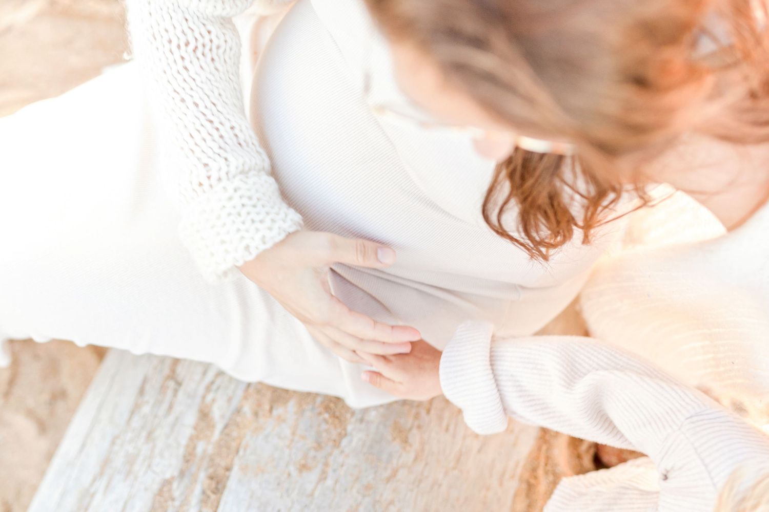 A side view of a pregnant woman in a white dress sitting on a beach.