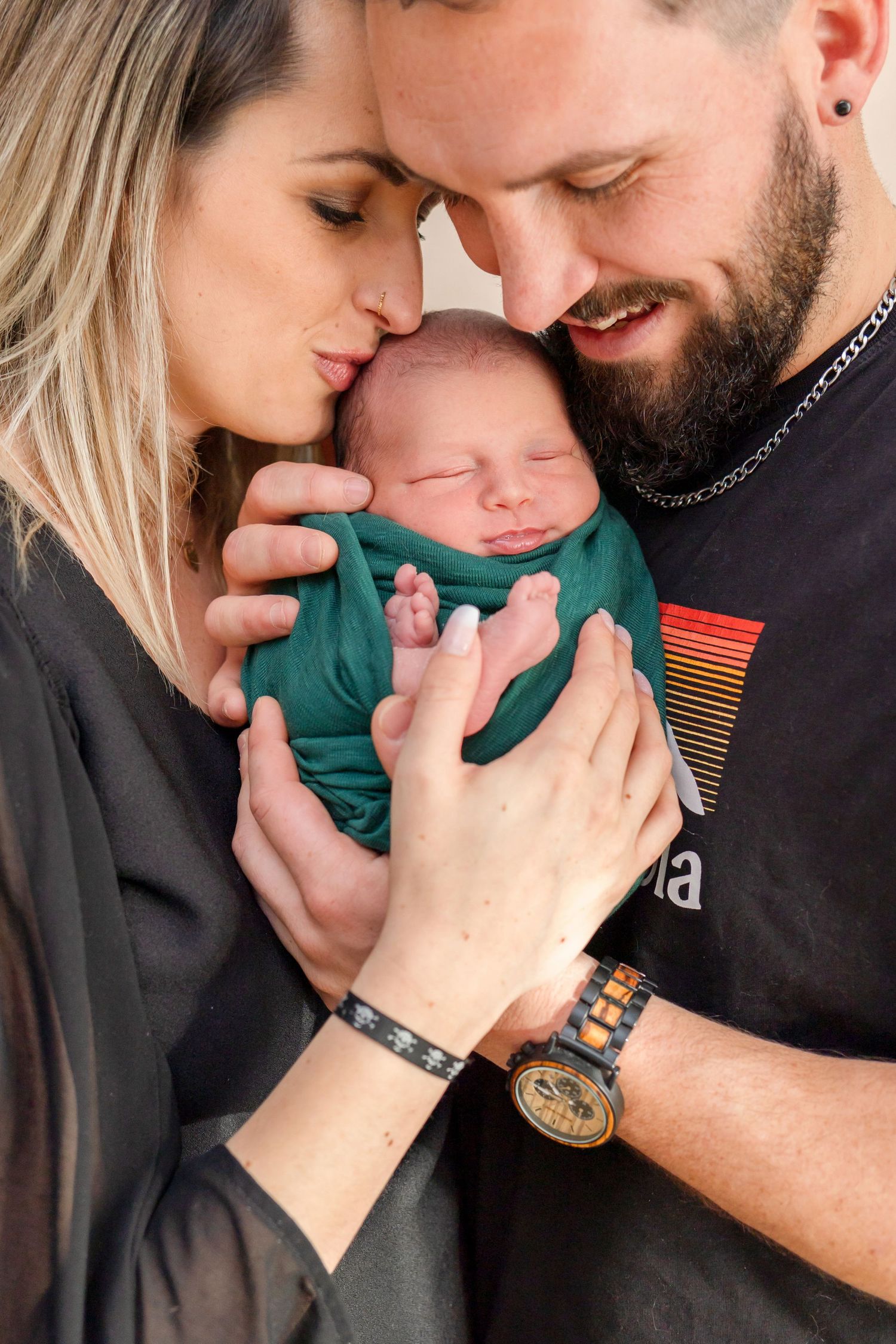 Tender moment as parents snuggle close to their sleeping newborn wrapped in teal fabric.