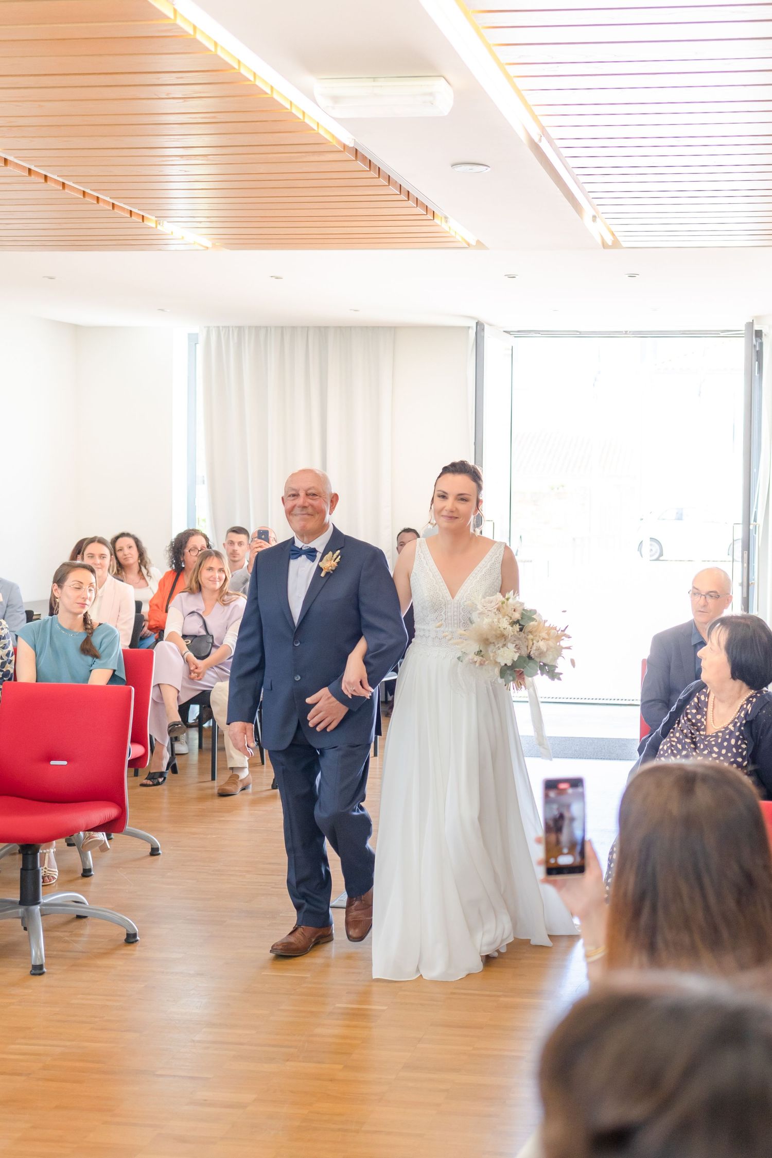 Wedding ceremony procession in bright indoor venue with wooden floors and red chairs.