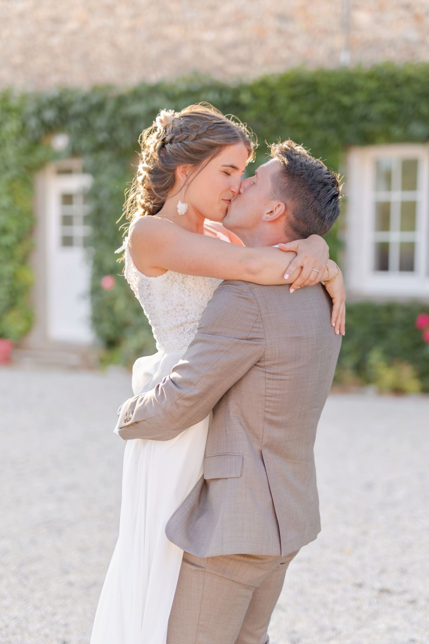 Romantic wedding portrait of a couple embracing outdoors against an ivy-covered stone wall in warm sunlight.