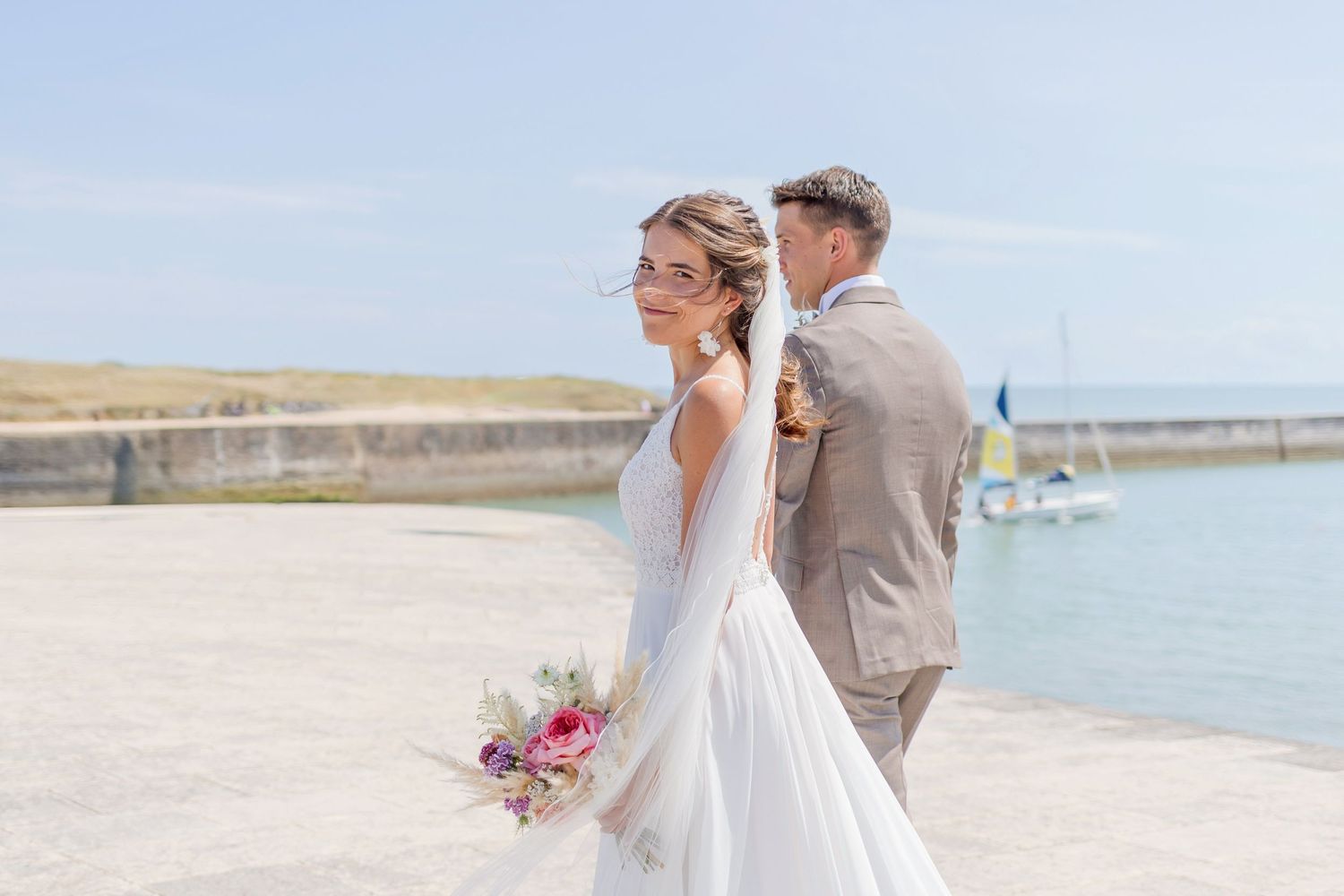 Wedding couple poses near lighthouse with ocean view and bouquet on bright sunny day.