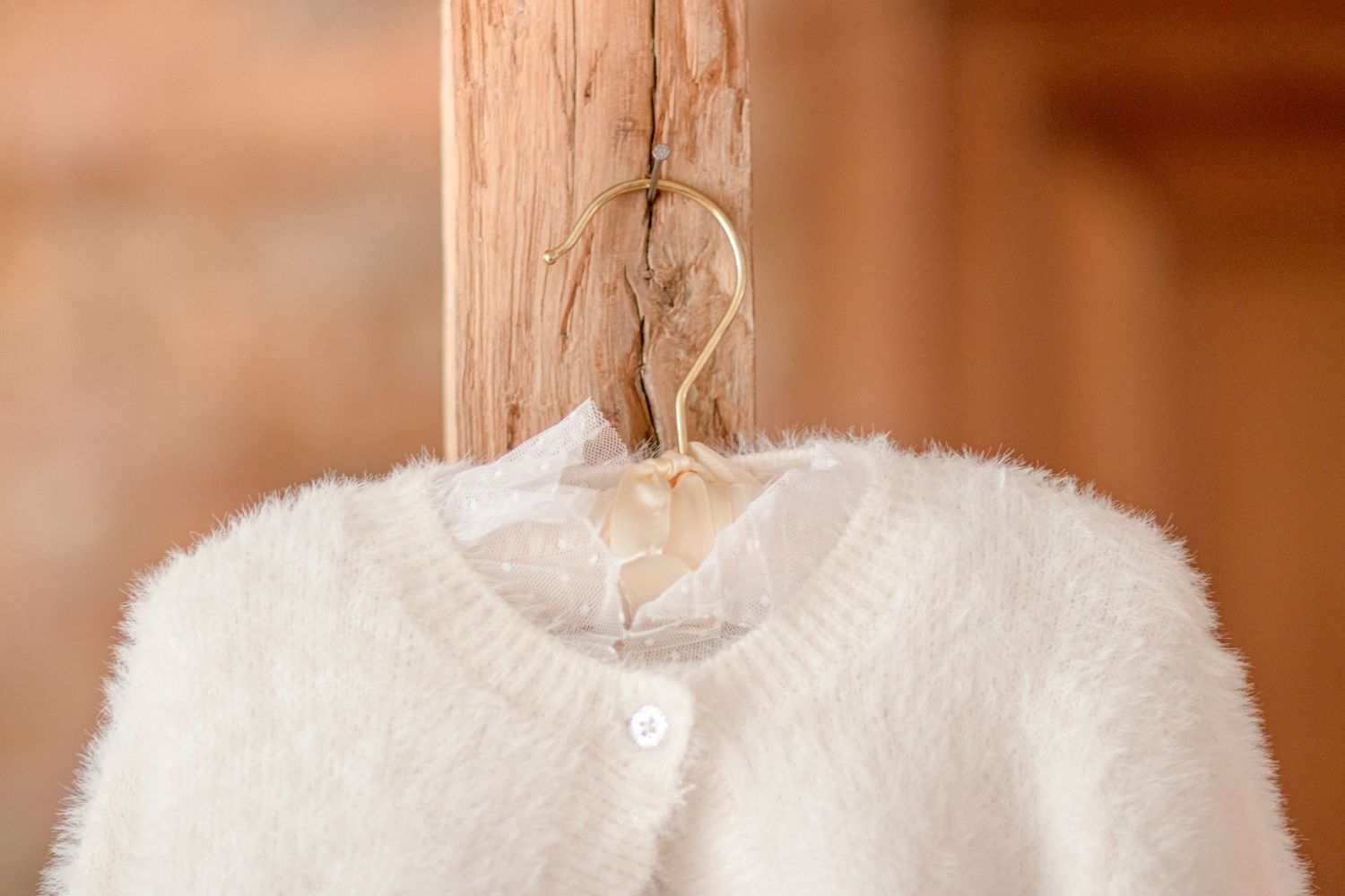 White fuzzy cardigan hanging on wooden beam against warm-toned background.