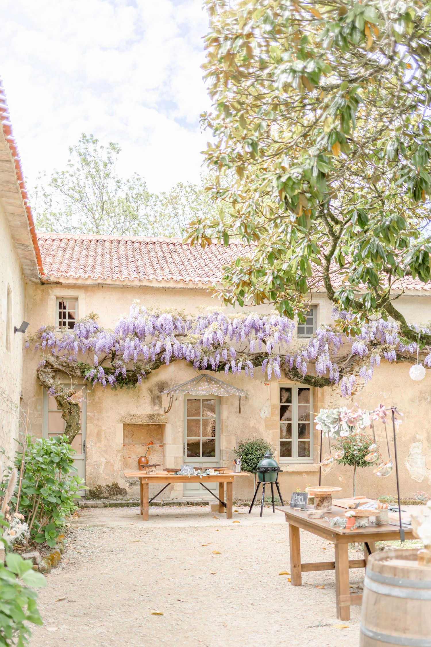 Charming stone cottage with blooming wisteria vines and wooden outdoor furniture in a courtyard.
