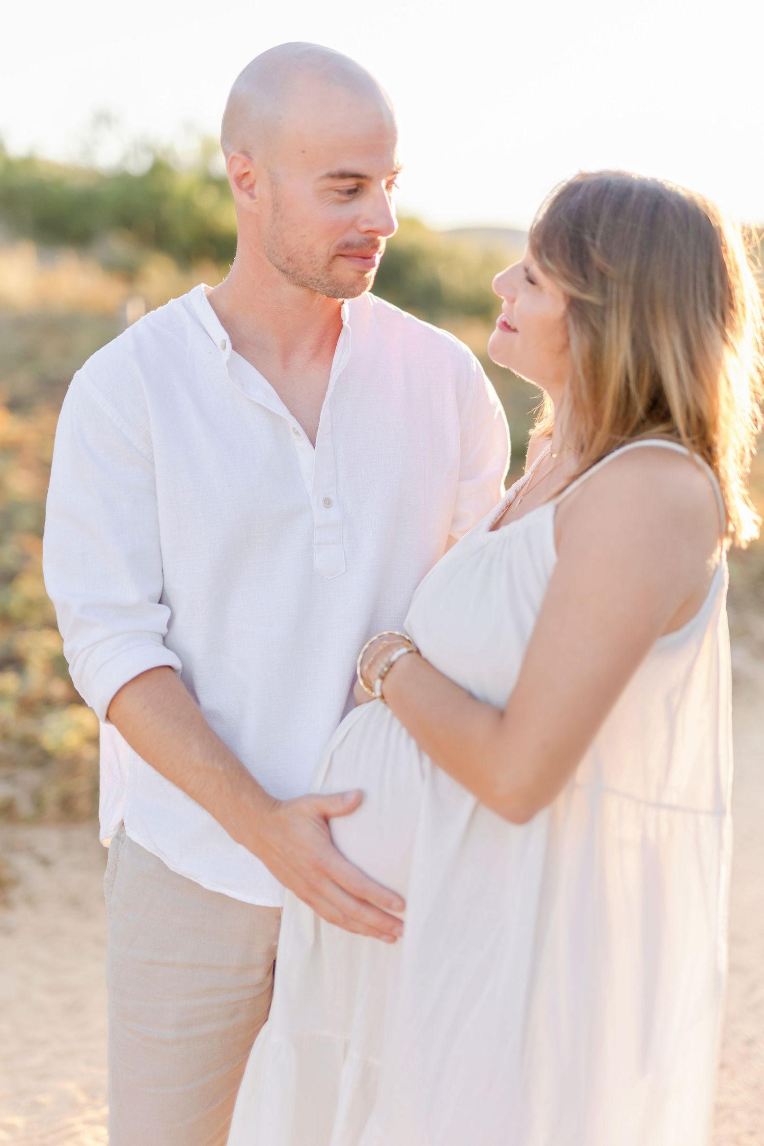 Couple in coordinating white outfits share tender moment during sunset maternity session.