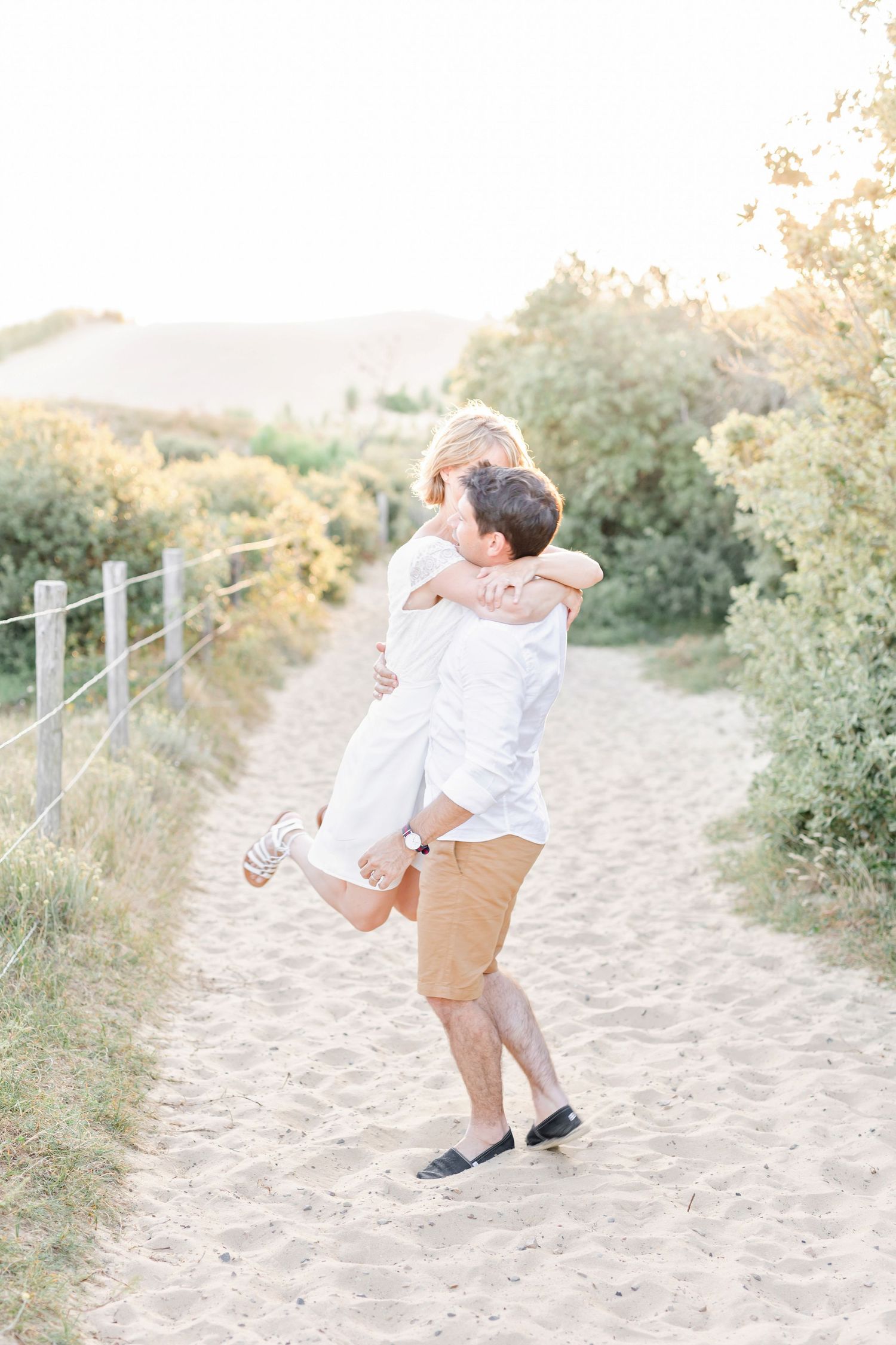 Playful couple dressed in white and khaki embraces on a sandy beach trail surrounded by coastal vegetation.
