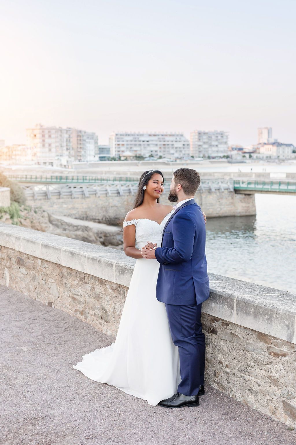 Bride in white gown and groom in navy suit share a tender moment on a seaside promenade.