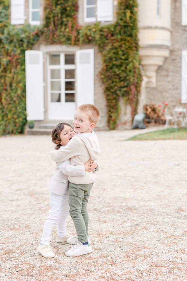 Two small children in light colored outfits play together near ivy-covered building.