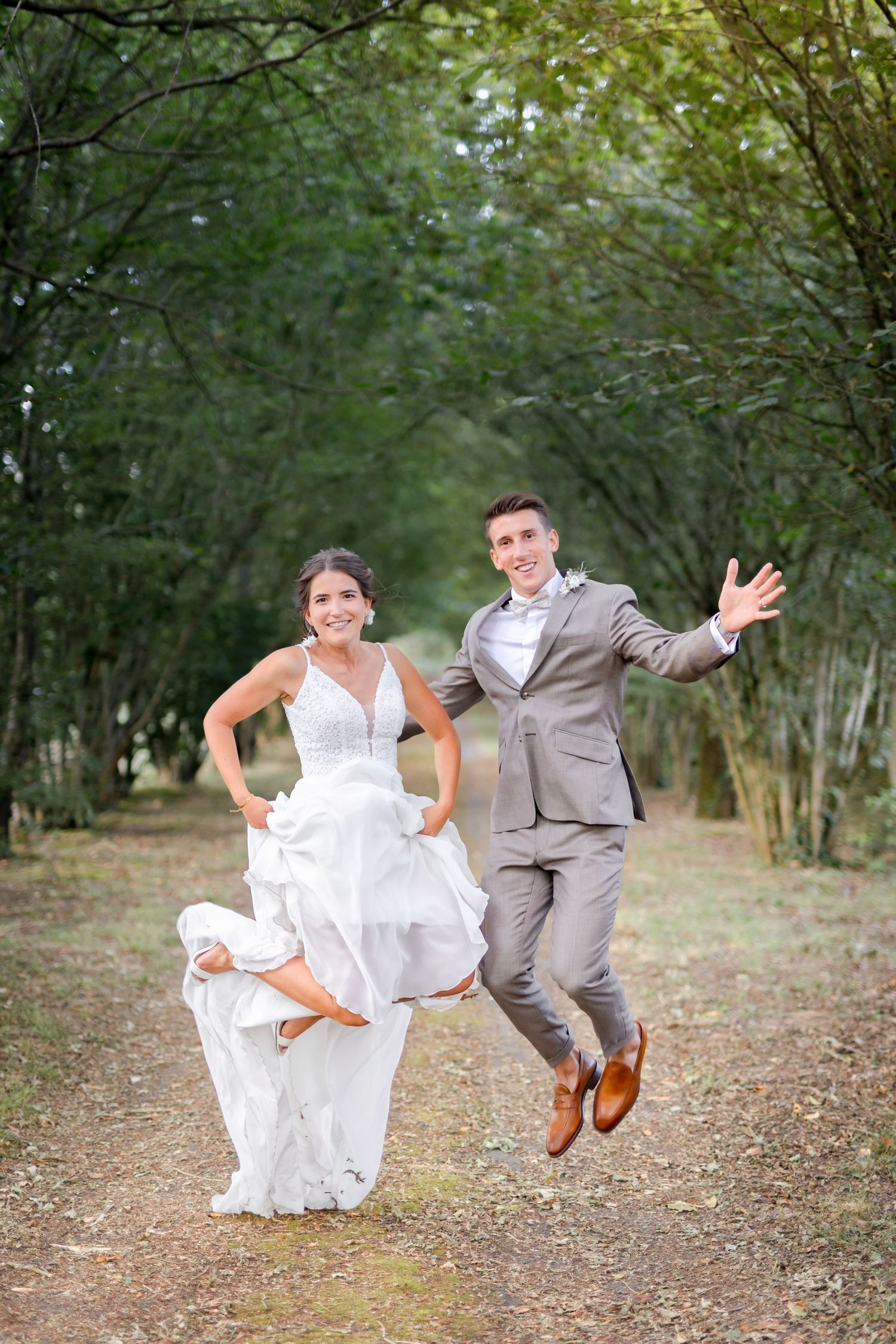 Newlyweds perform fun jumping poses for photos on a tree-lined path.