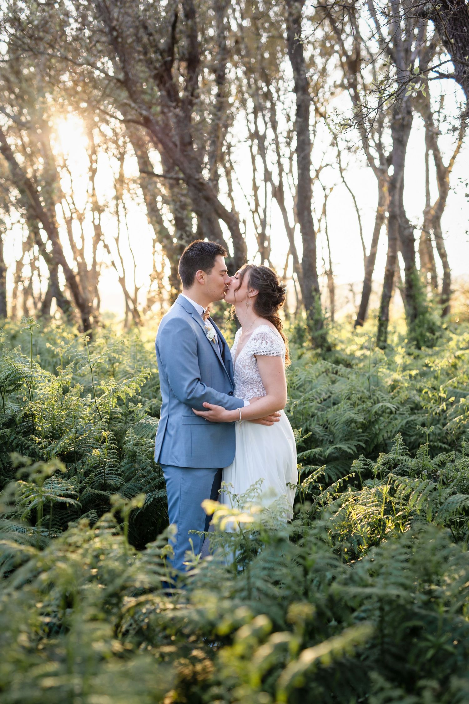 Newlyweds share a romantic moment in a sunlit forest surrounded by lush green ferns.