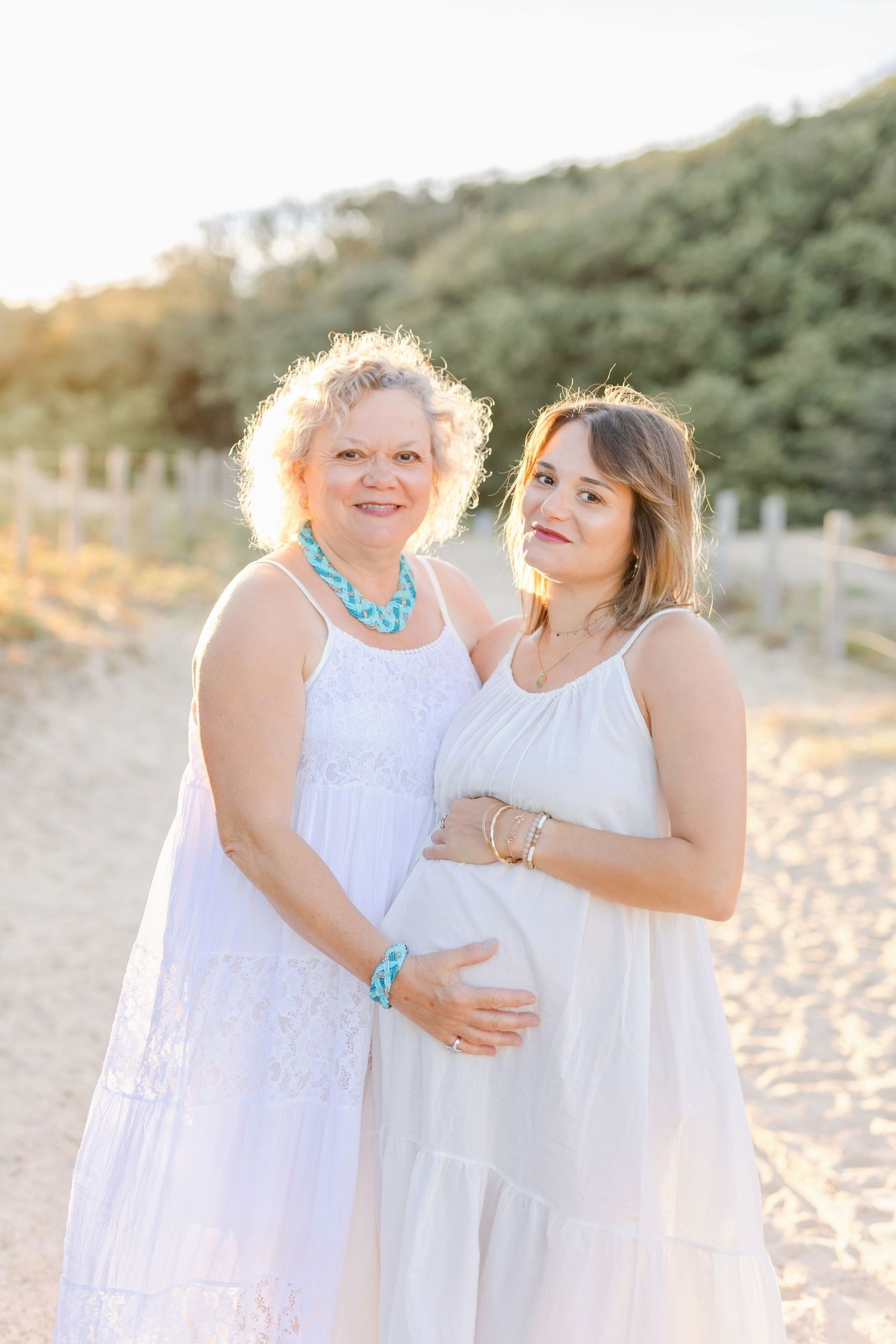 Three women in white dresses share joyful moment during beach photoshoot at sunset.