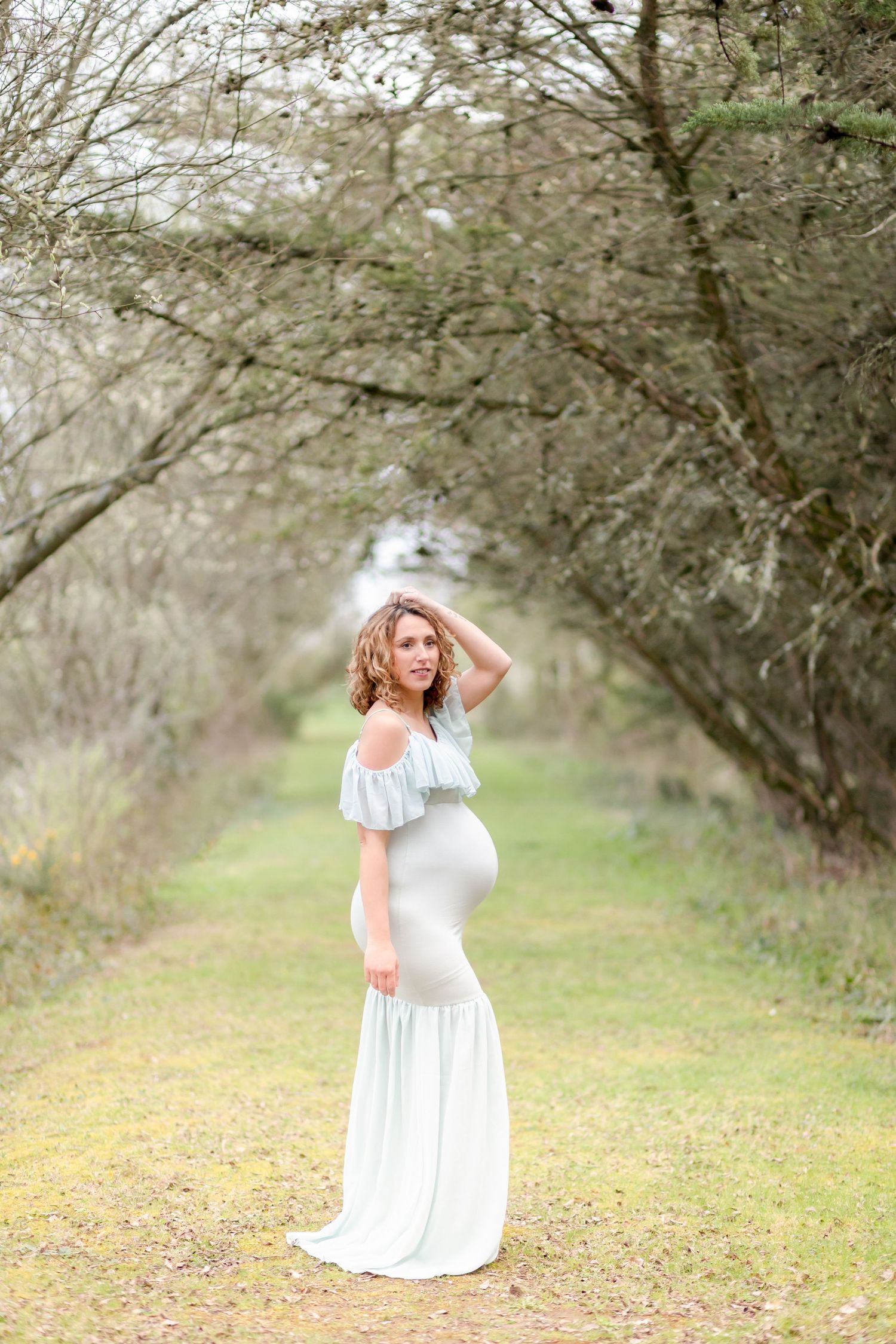 A elegant maternity pose captured in a natural tree-lined pathway with soft lighting.