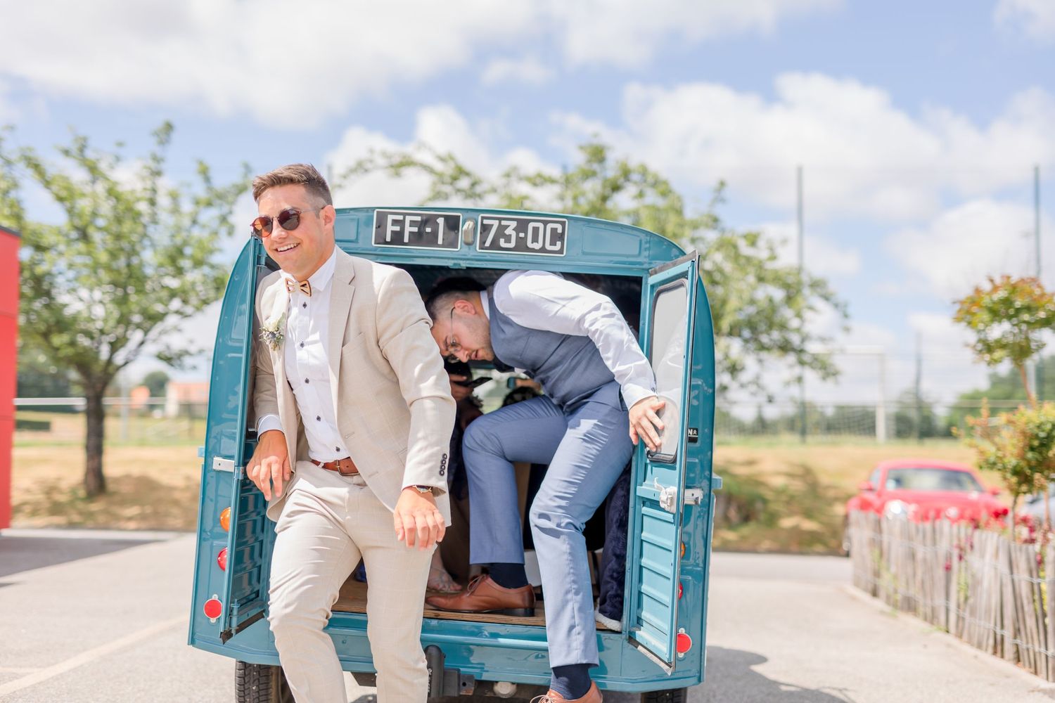 Wedding guests pose playfully from back of vintage blue Citroen van on sunny day.