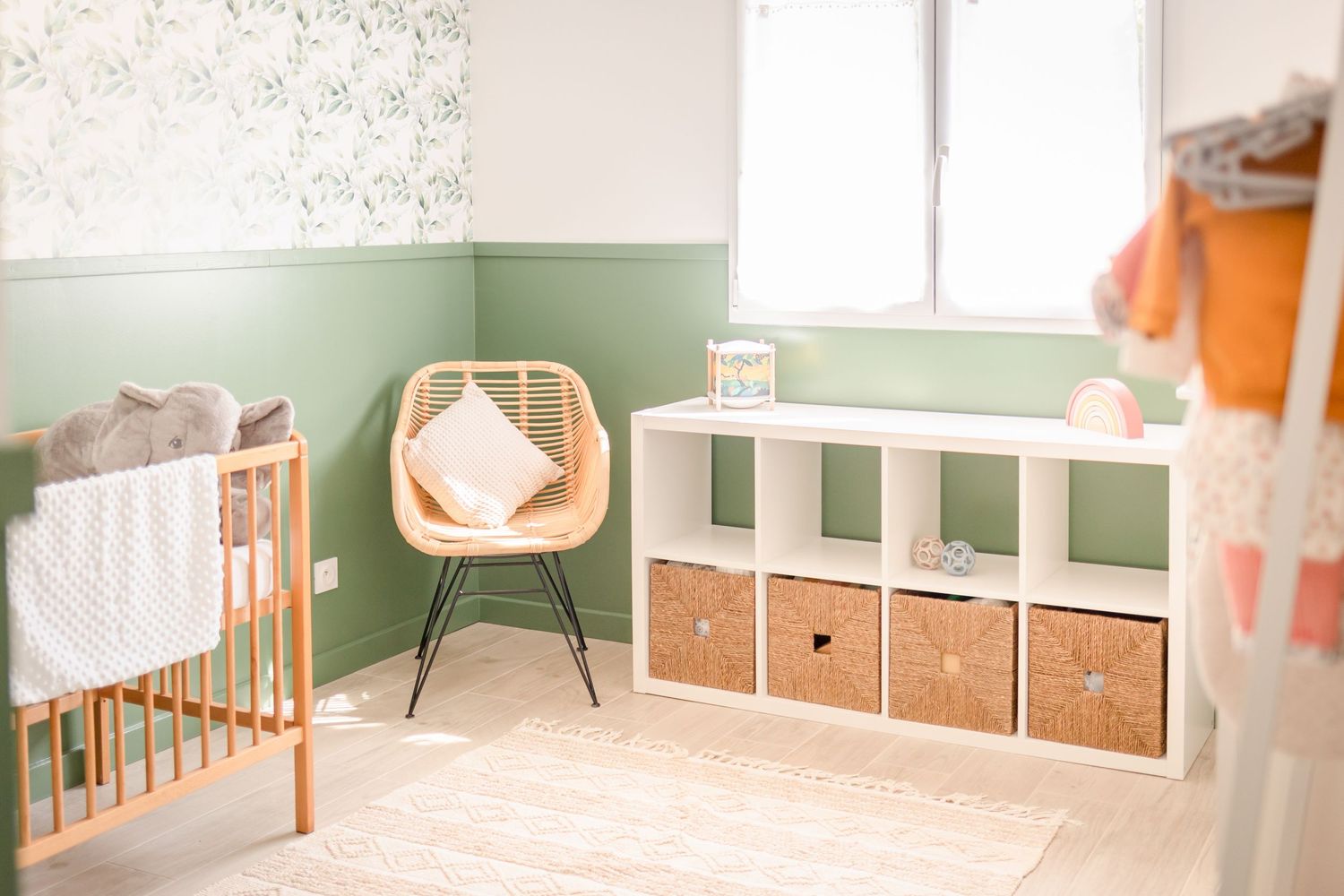 Modern nursery with sage green walls, rattan chair, and white storage unit featuring natural baskets.