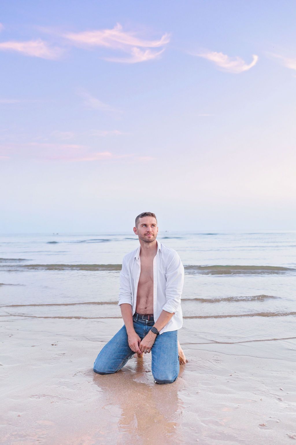 A person in a white shirt and jeans kneels on the sandy beach at sunset with ocean waves in the background.