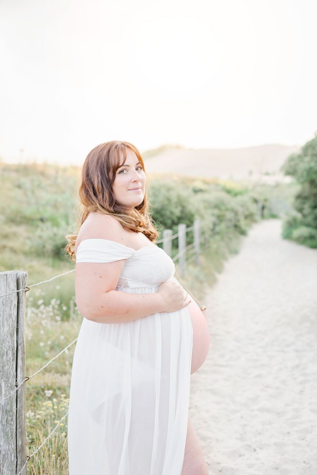 Woman in flowing white dress poses along sandy beach path with coastal vegetation.