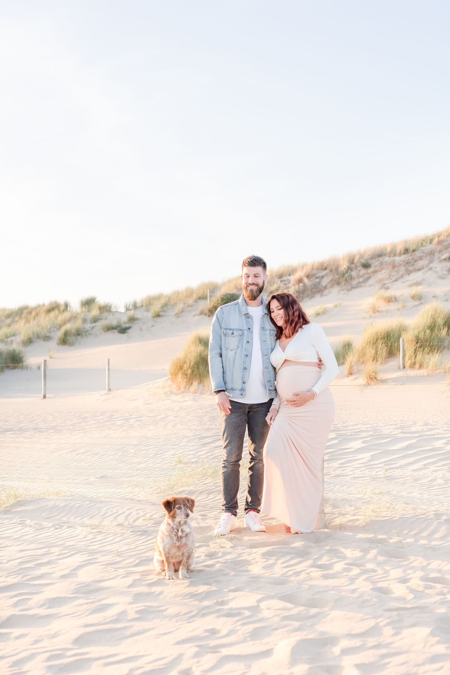 A couple walks along a sandy beach with their small dog during golden hour.