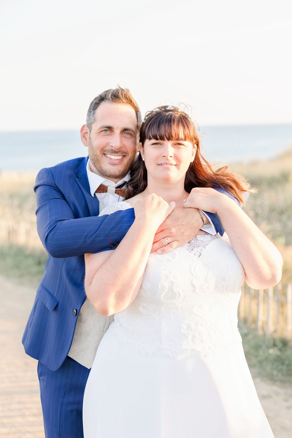 A joyful wedding couple embraces in front of a coastal lighthouse on a sunny day.