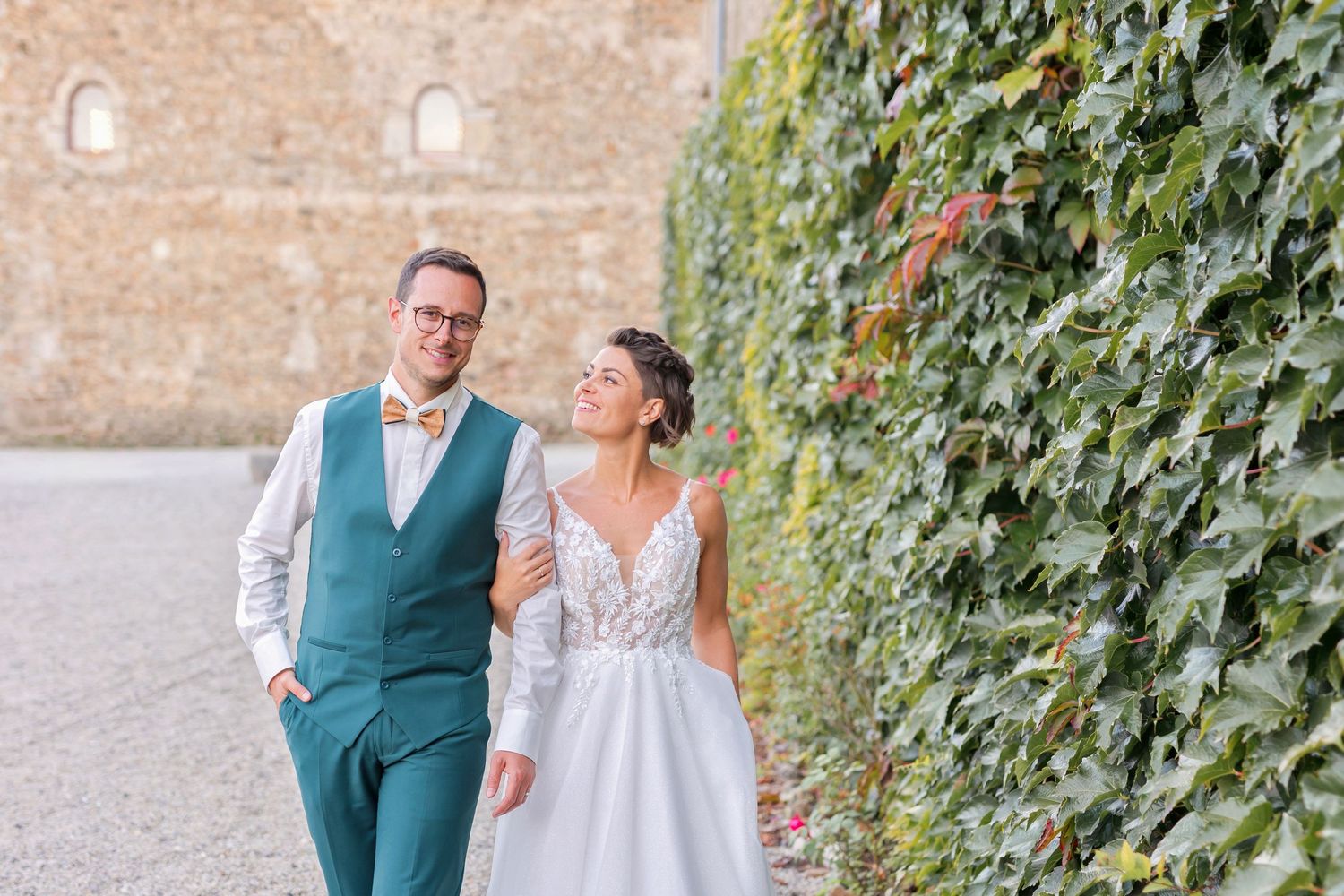 A couple walks along a brick wall with ivy, the bride in a lace gown and groom in a teal vest.