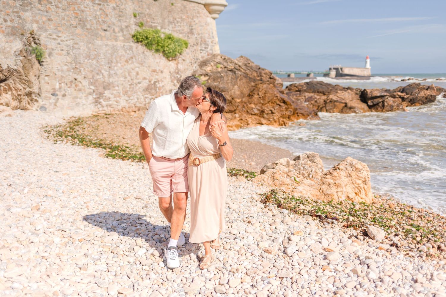 Casual beach stroll along stone fortress wall with lighthouse visible in distance.
