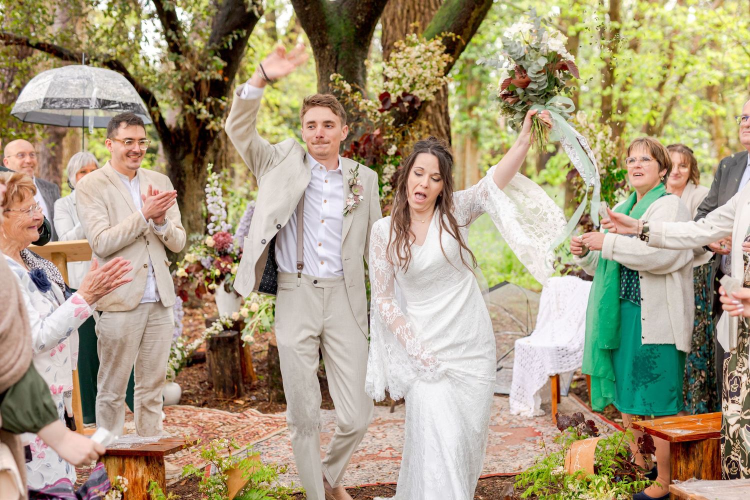 Jubilant newlyweds raise their arms in celebration while guests applaud at a woodland ceremony.