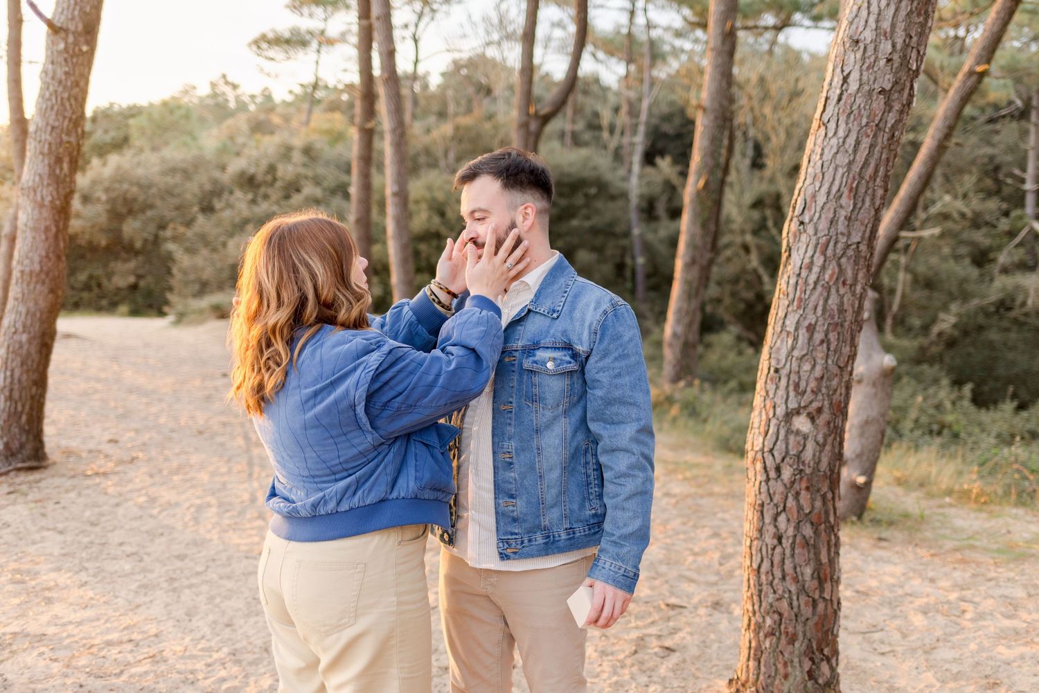 A couple wearing denim jackets shares a playful moment in a pine forest.