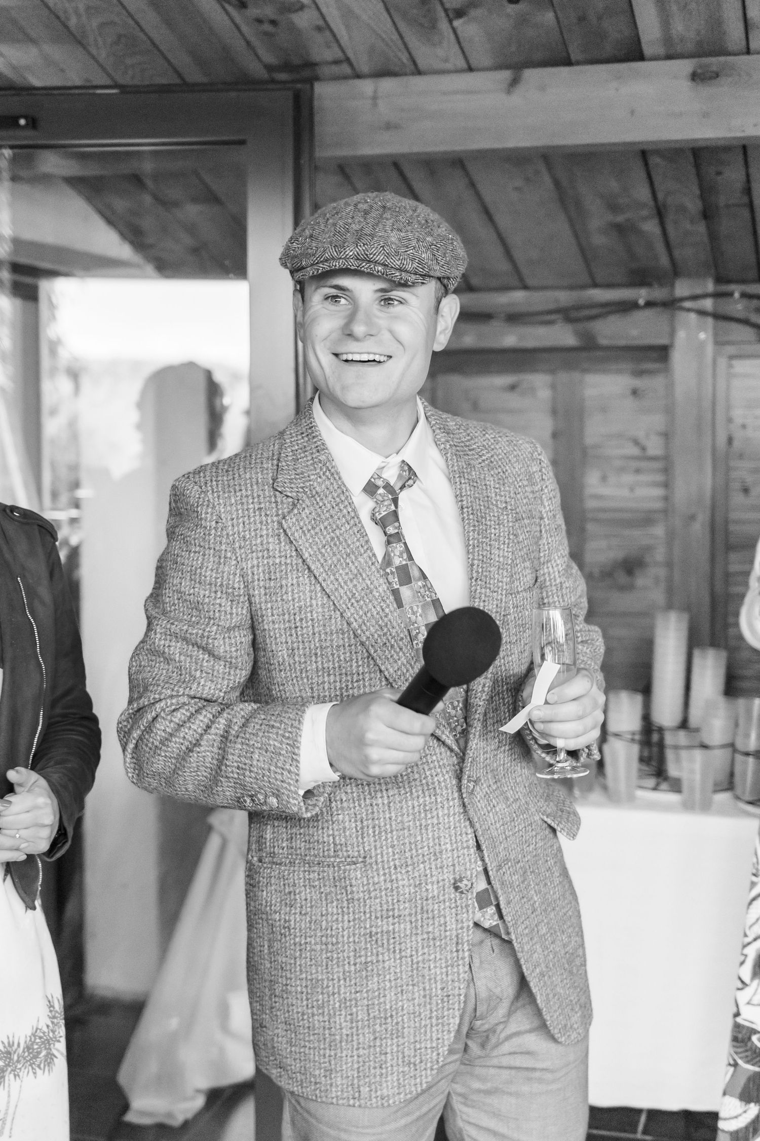 Wedding guest in tweed suit and flat cap holding microphone indoors.