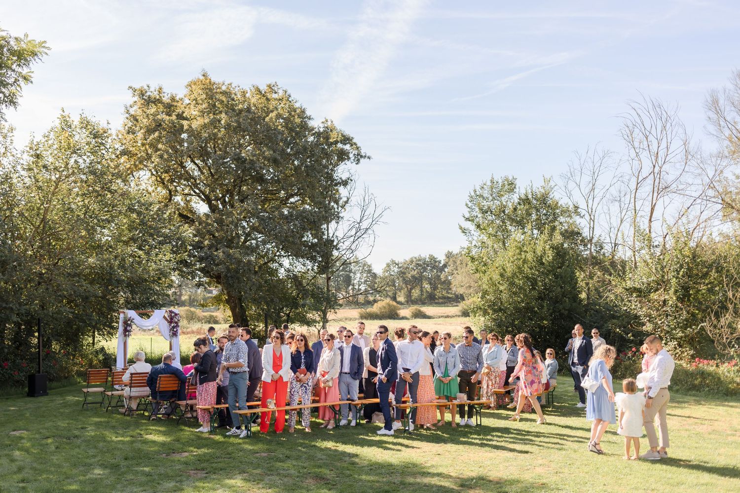Wedding guests gather outdoors for a ceremony under trees on a sunny day.