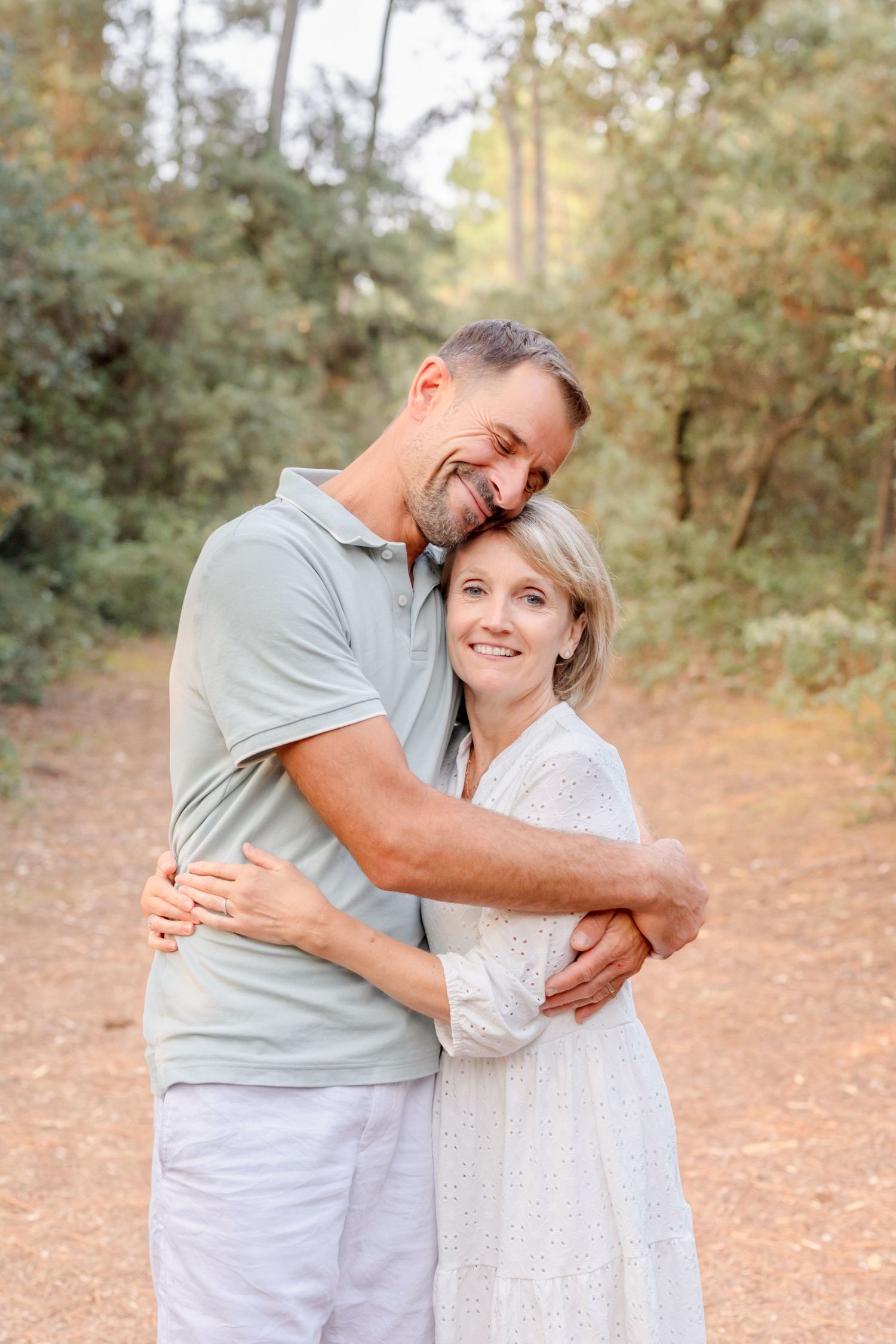 Two people in light colored clothing share an affectionate moment on a sunlit forest trail.