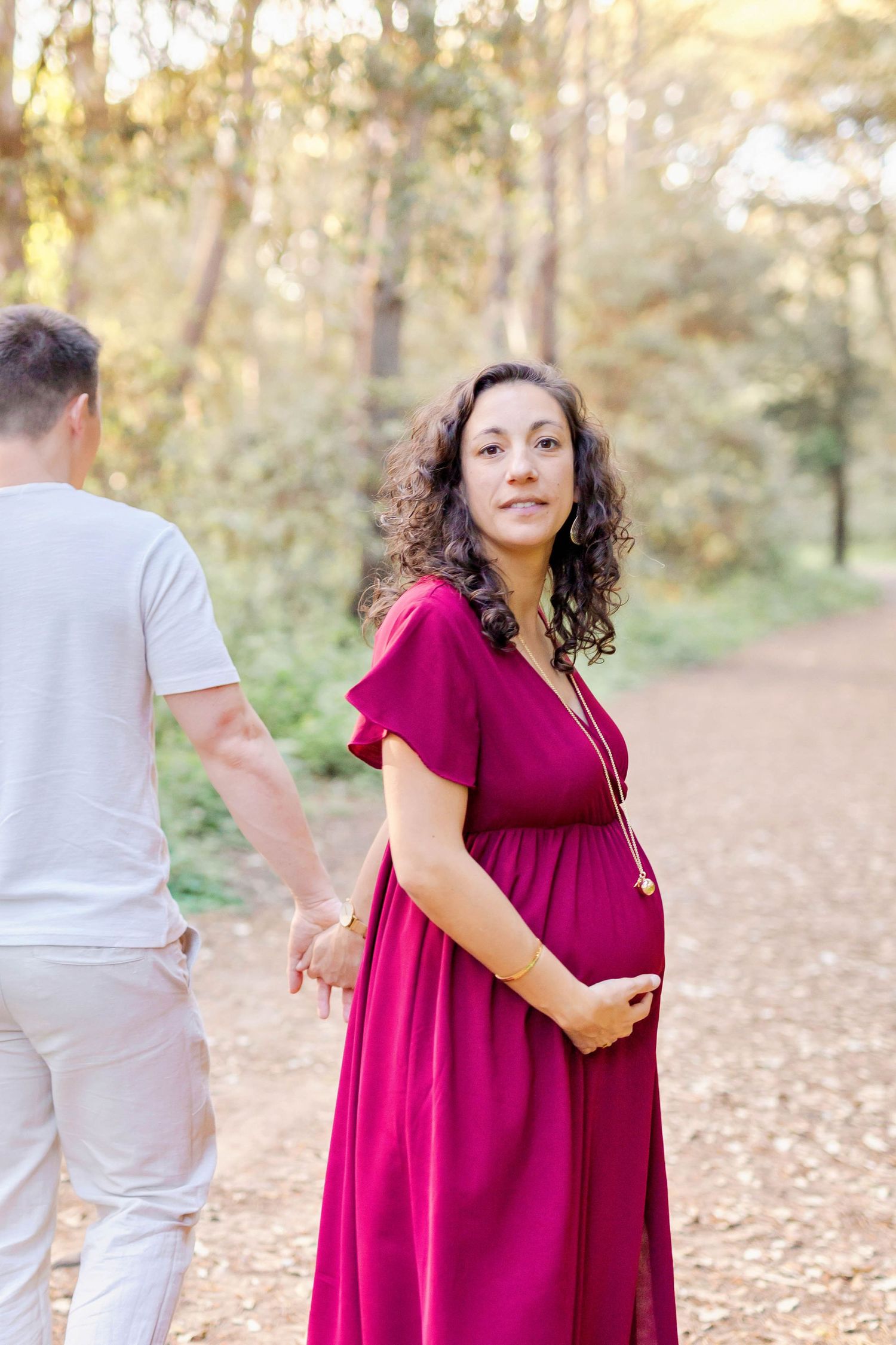 A maternity session features tender moments captured on a leaf-covered trail in golden sunlight.