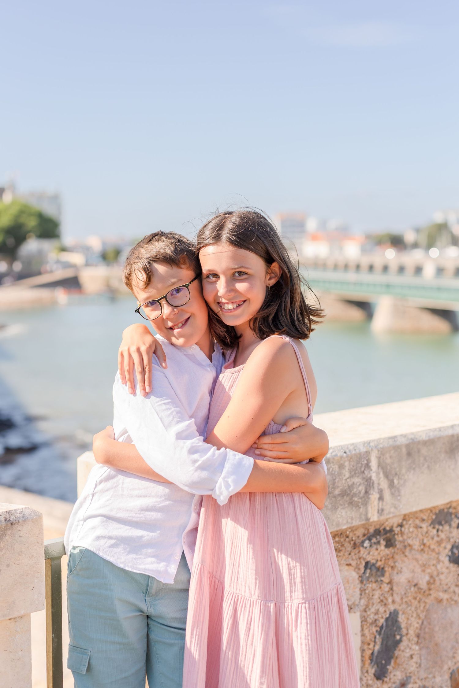 Two people embracing by waterfront with bridge and city skyline in background.