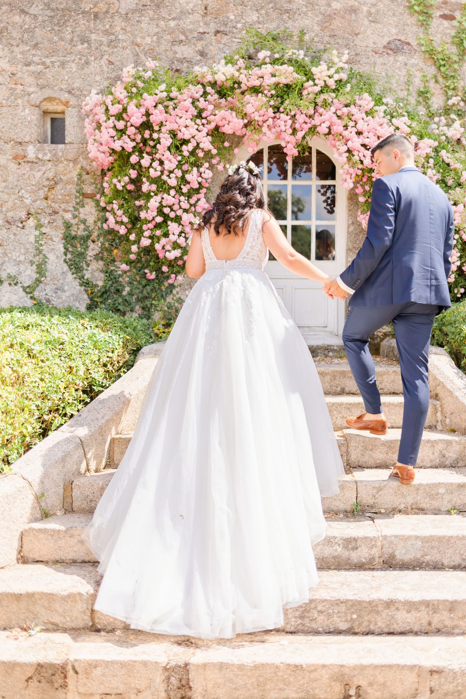 Elegant couple in wedding attire walking along stone steps with pink flowering bushes.