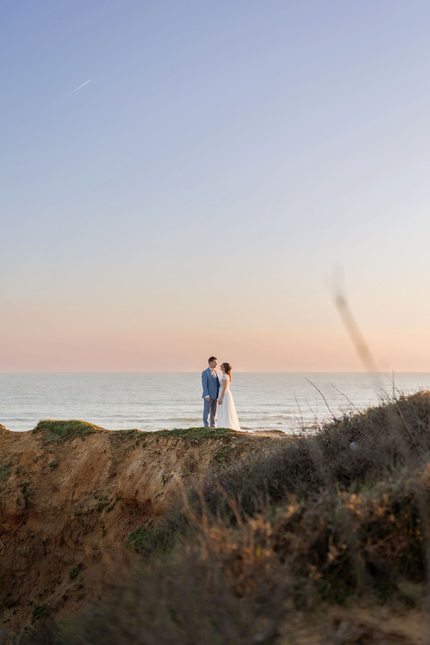 Wedding couple standing on coastal cliffs overlooking ocean at golden hour.