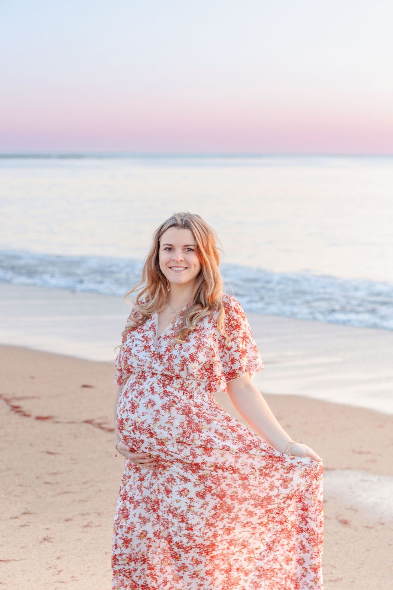 Woman in flowing red floral dress twirling on beach at sunset.