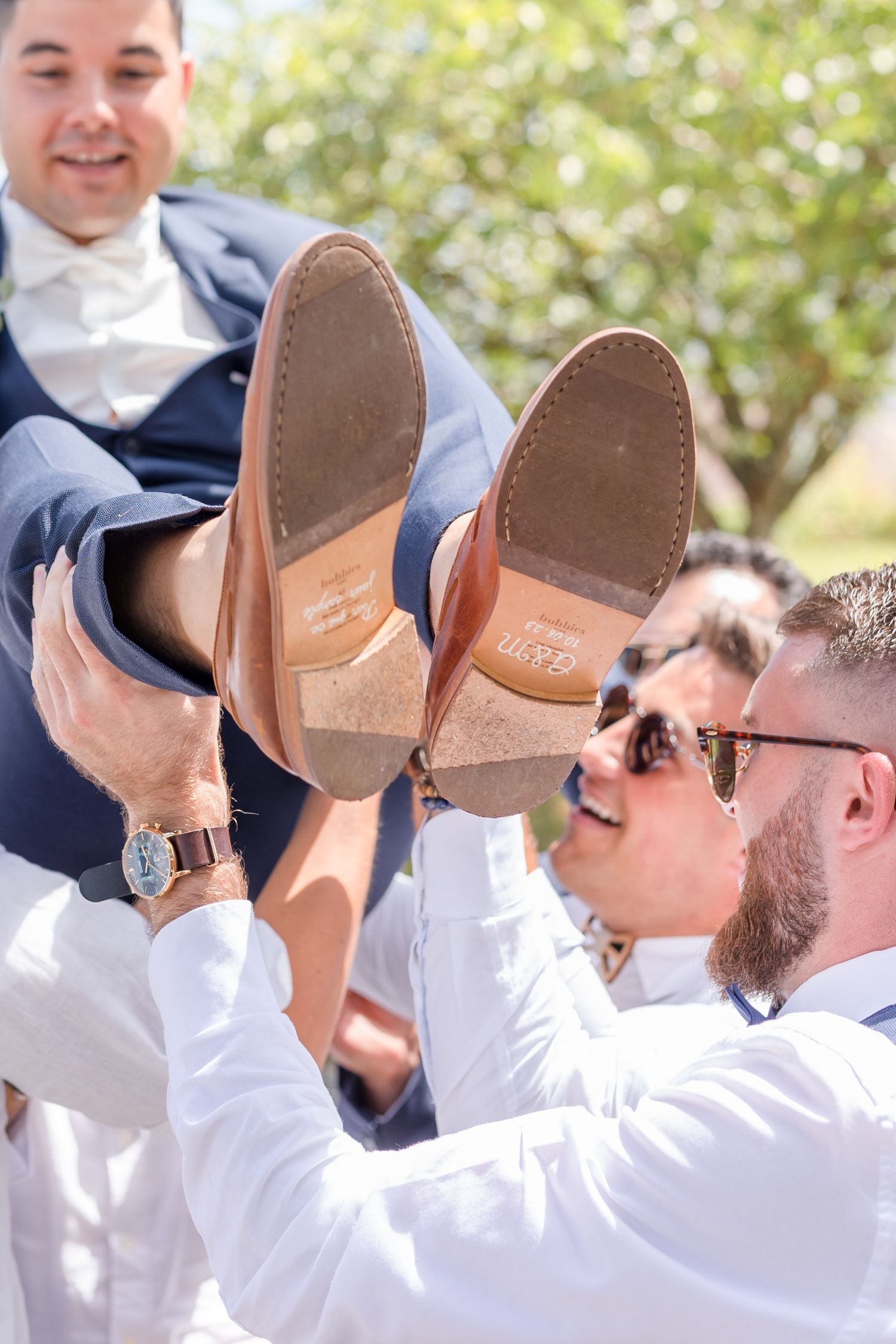 Wedding party members holding up dress shoes while posing outdoors.