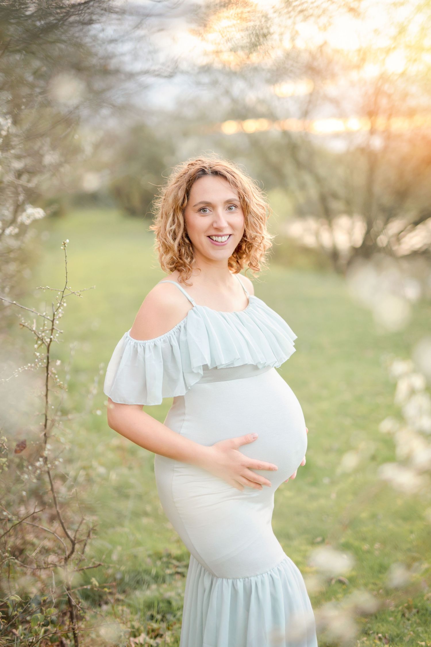 Ethereal maternity portrait in a light blue dress with ruffle details against a soft sunset background in nature.