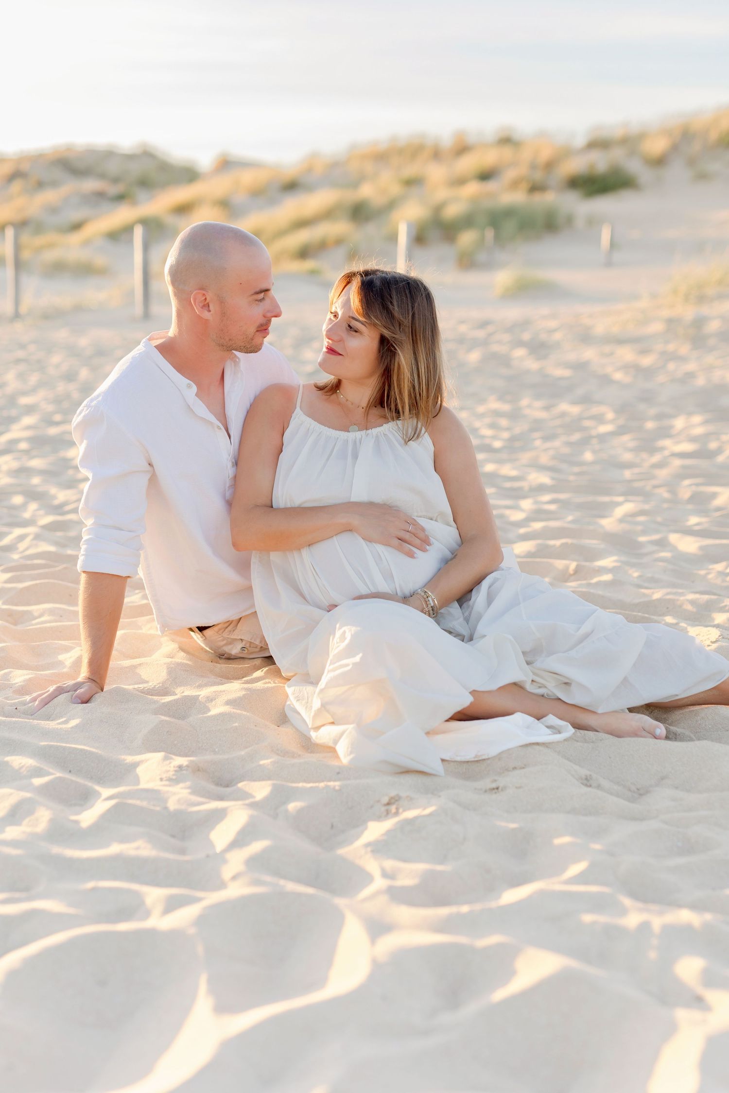 A couple in white outfits shares an intimate moment on a sandy beach at sunset.