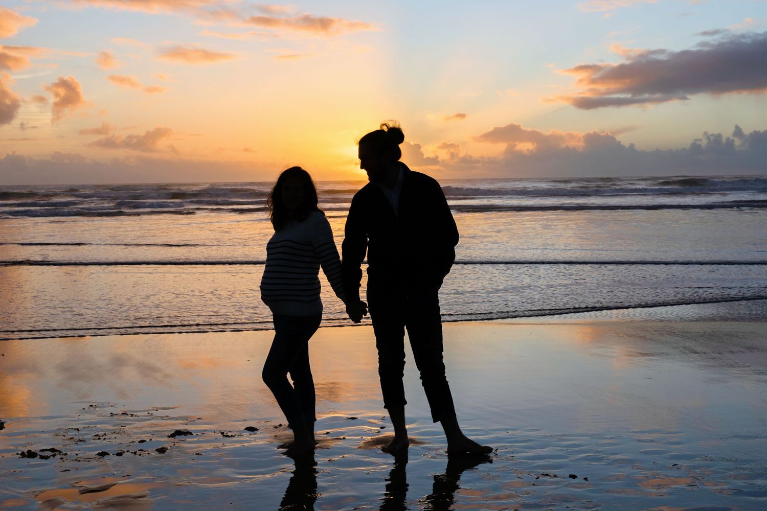 Two silhouettes stand hand in hand on a beach at sunset.