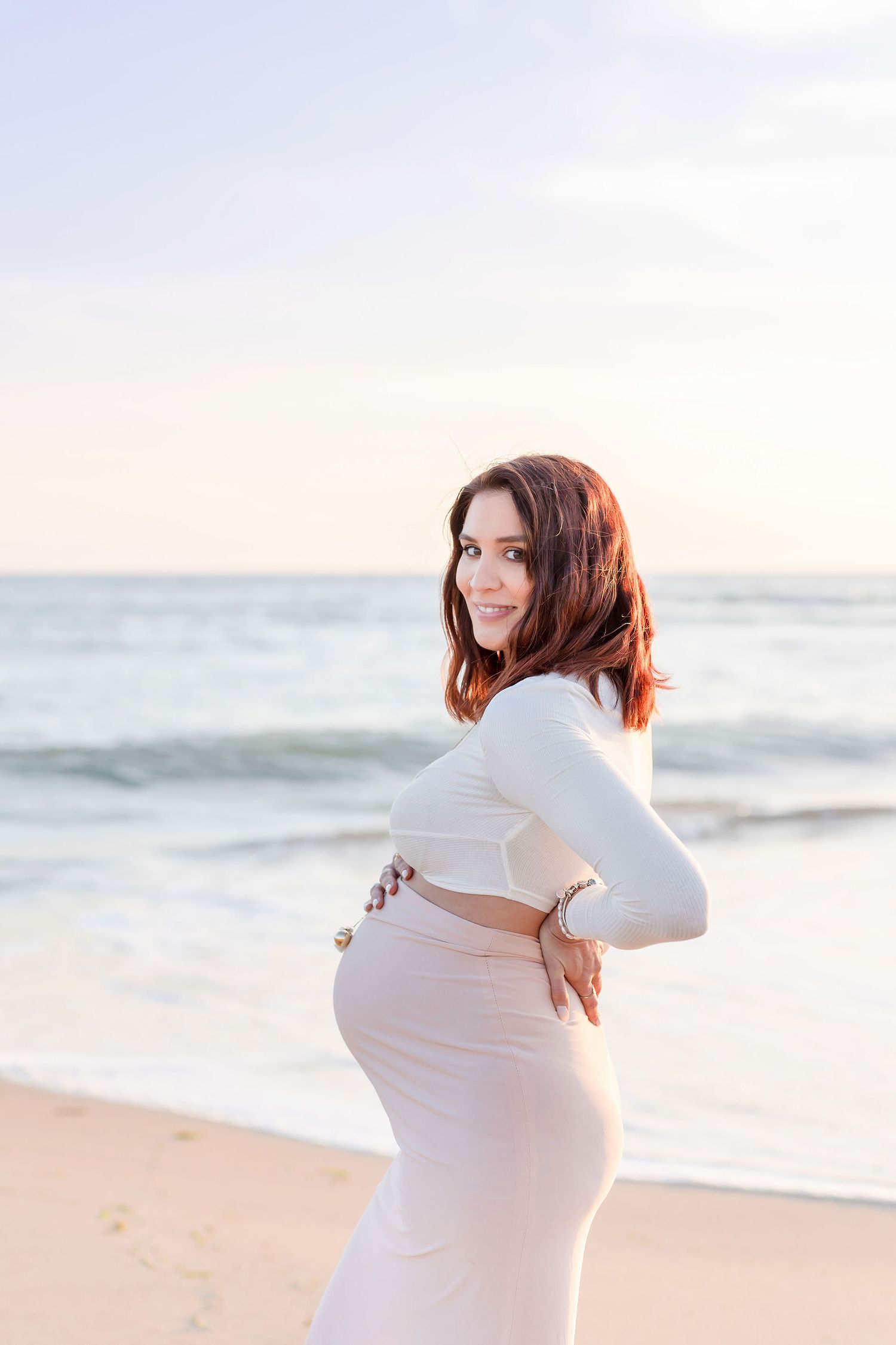 A maternity photo shoot on the beach shows a woman in white embracing her baby bump.
