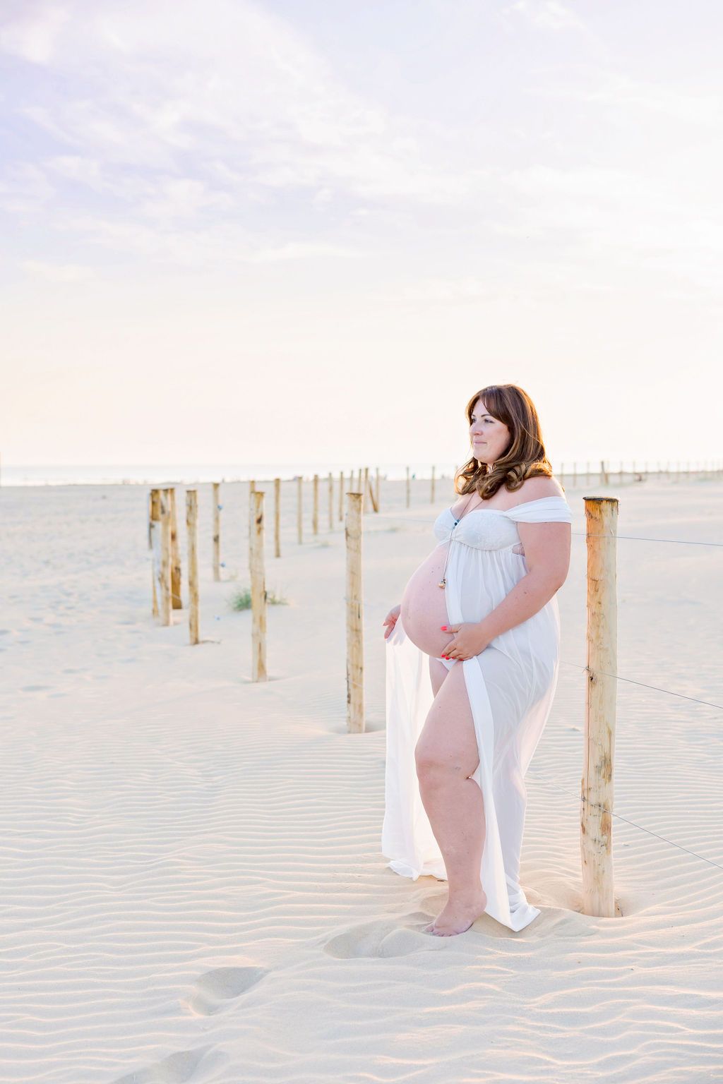 Expectant mother in white dress posed by wooden posts on sandy beach at sunset.