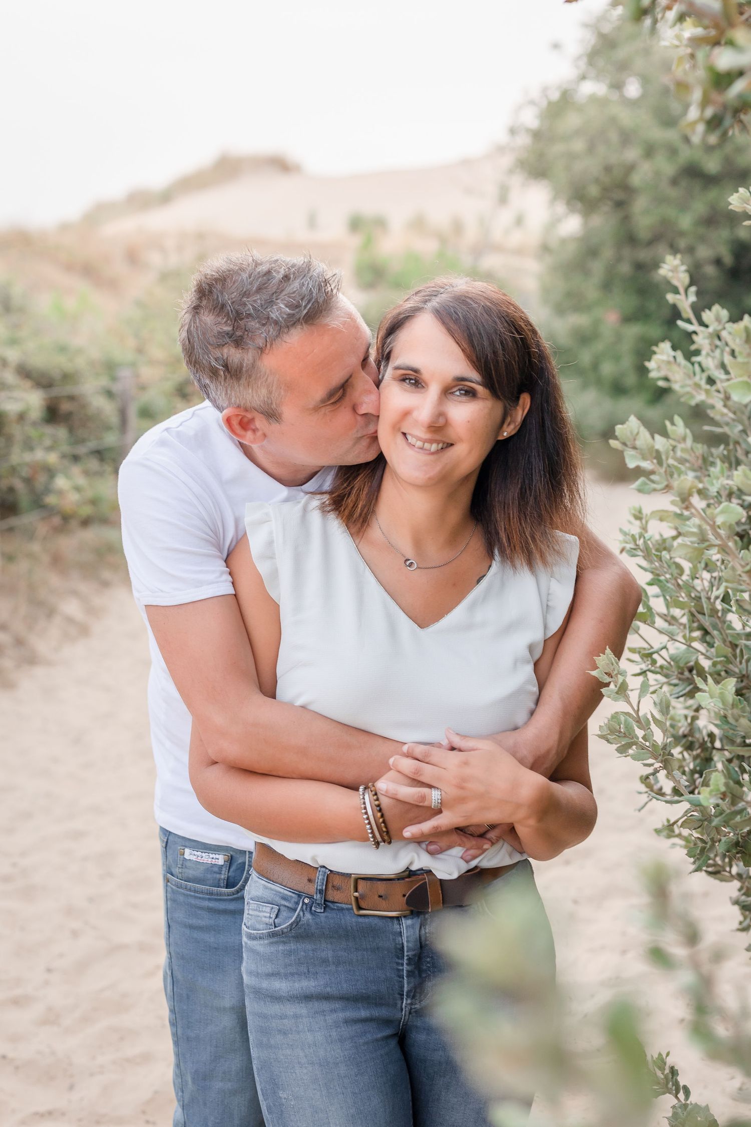 Romantic couple embracing on sandy trail surrounded by desert brush during golden hour photo session.