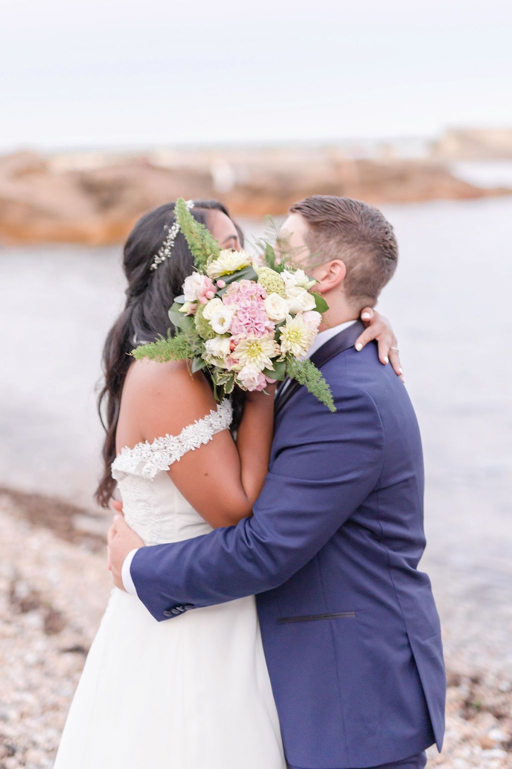 A romantic wedding moment captured as a couple embraces on a rocky beach.