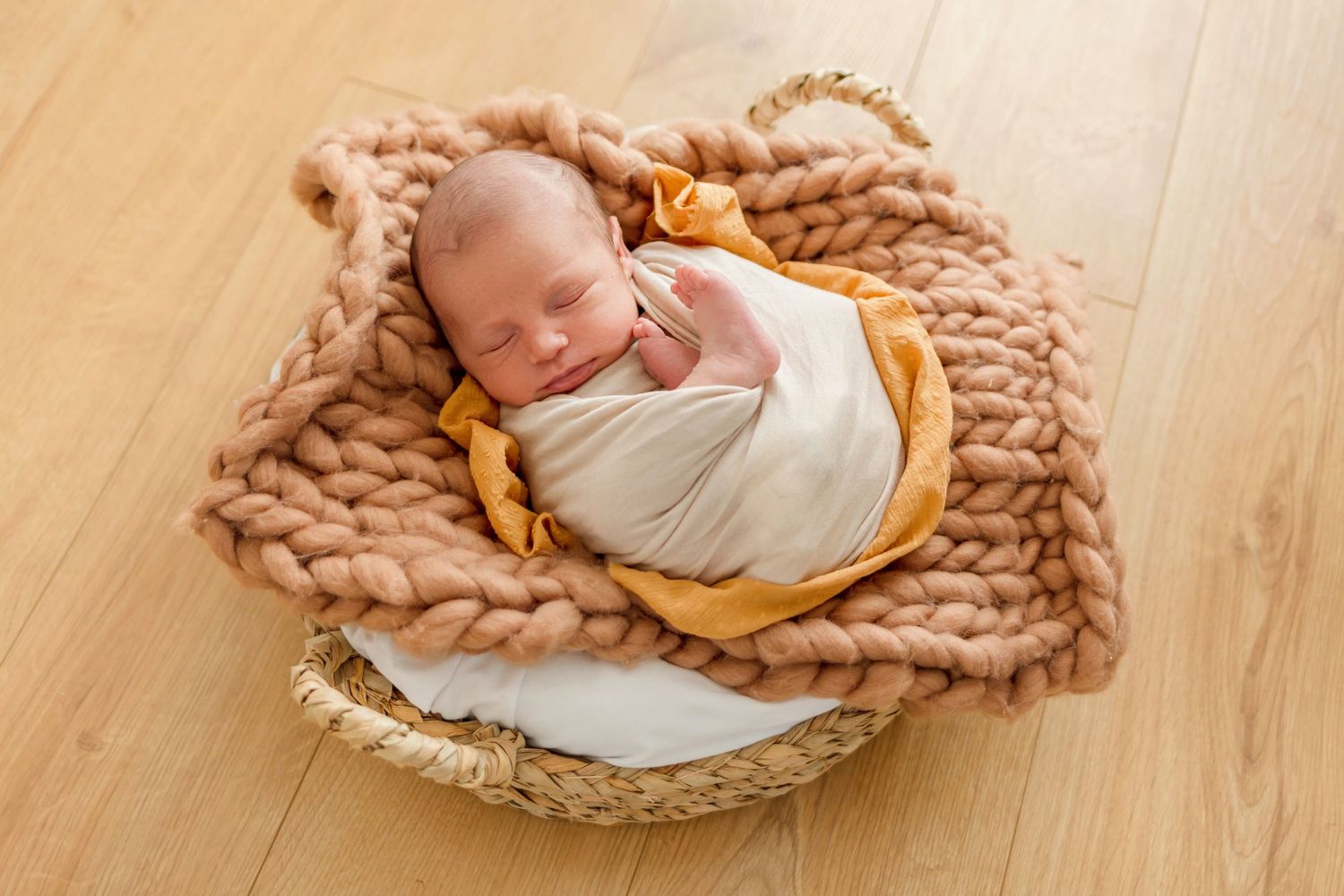 Sleeping newborn swaddled in white and yellow wraps nestled in chunky knit blanket on wooden floor.