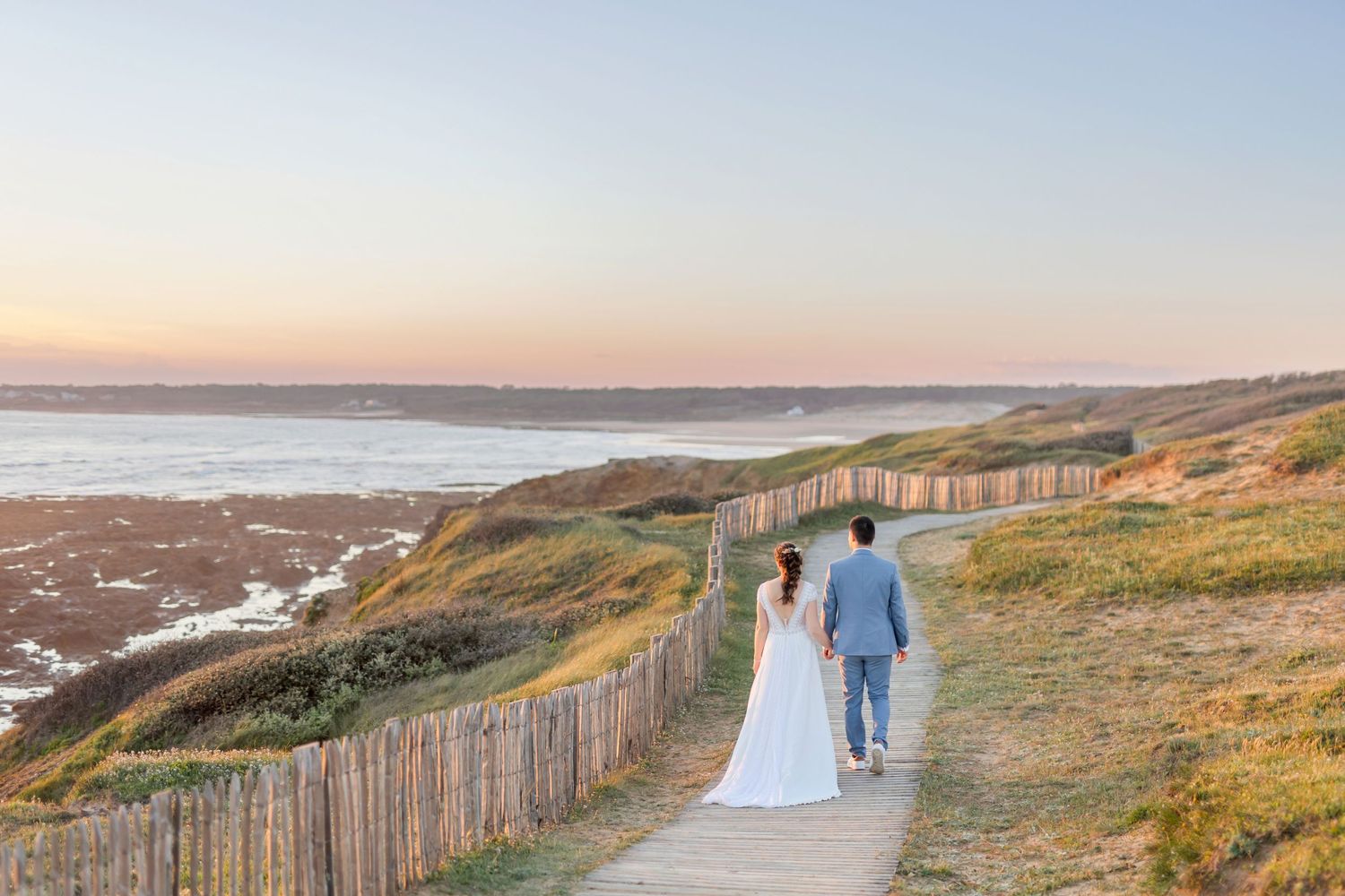 Newlyweds walking along wooden boardwalk path by coastal cliffs at sunset.