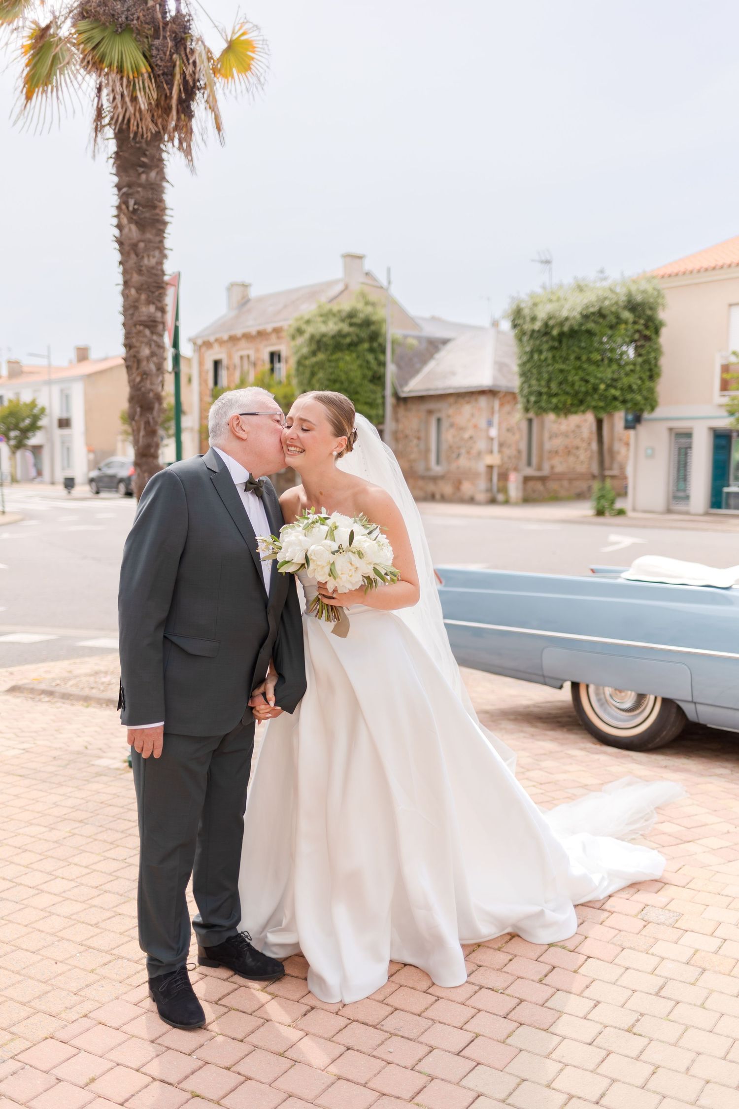 A couple shares a tender moment by a vintage car on their wedding day with a palm tree in the background.