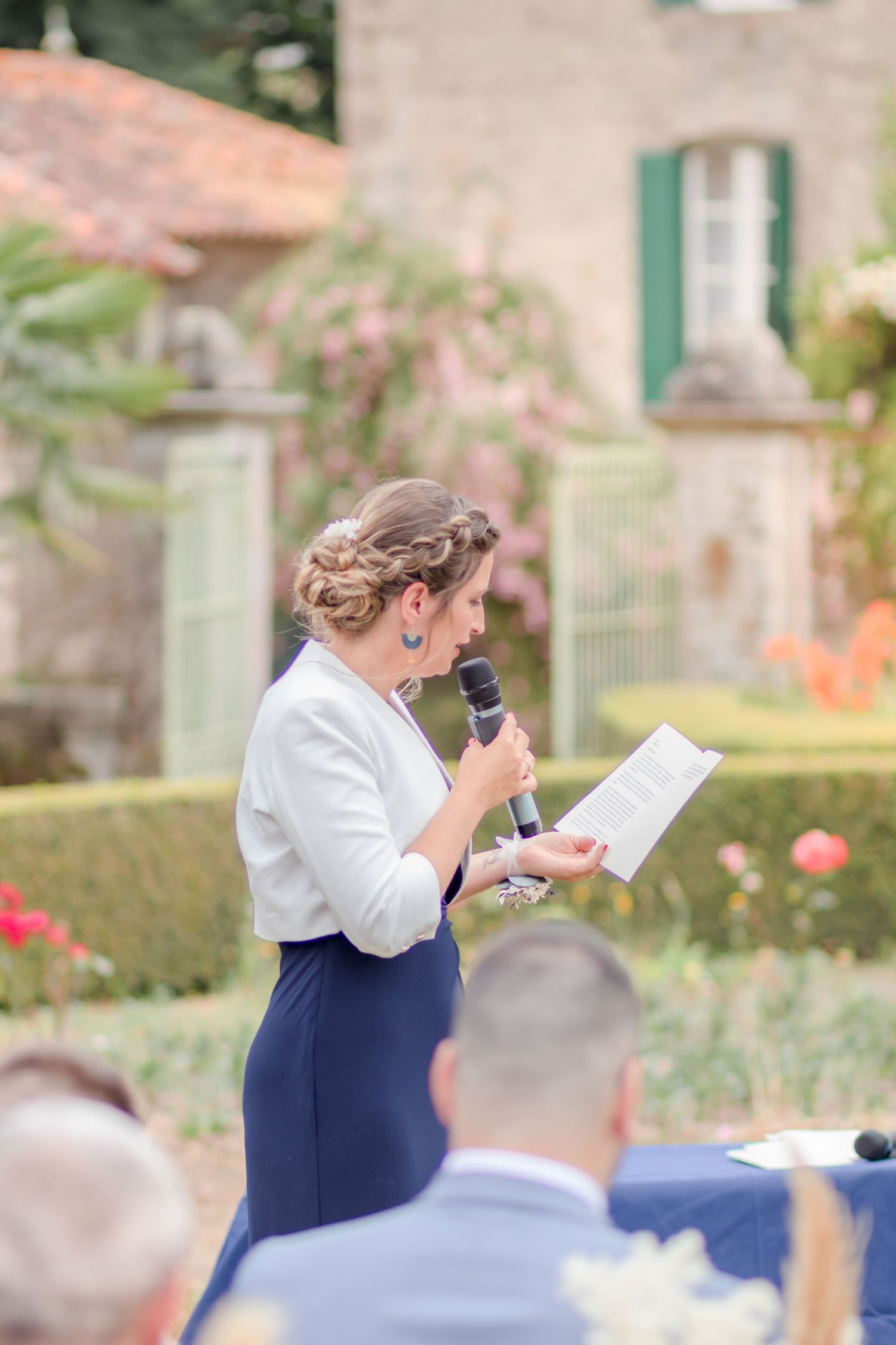 A woman in formal attire gives a speech at an outdoor event with a microphone.
