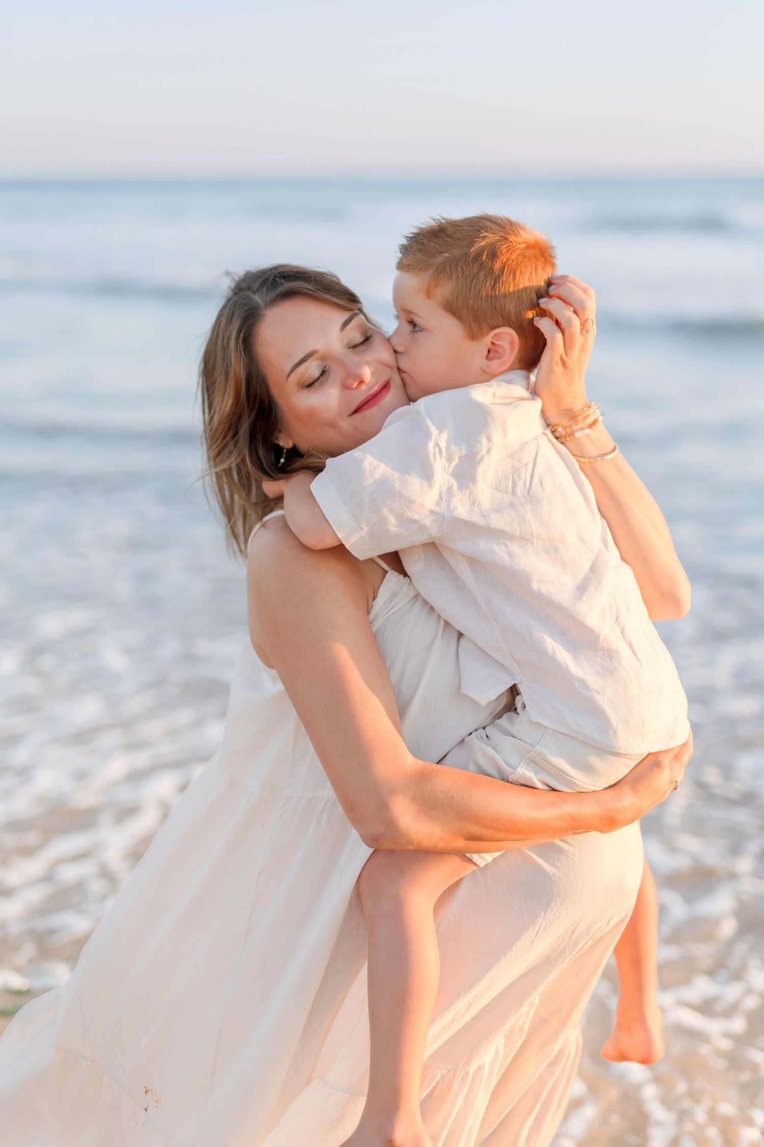 Tender moment captured between figures in white clothing on beach with waves in background.