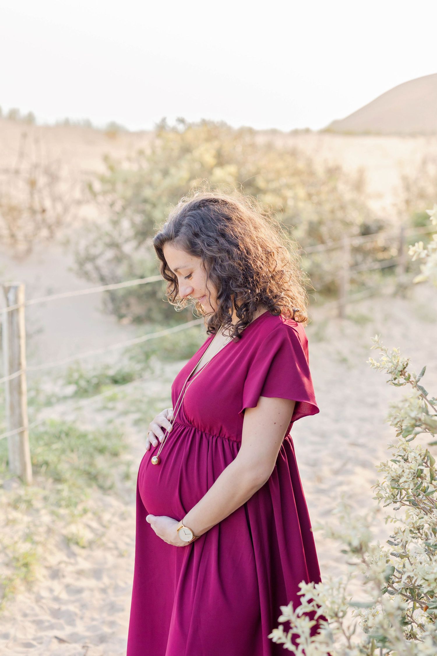 Side profile maternity photos in a desert setting with woman wearing flowing burgundy dress.