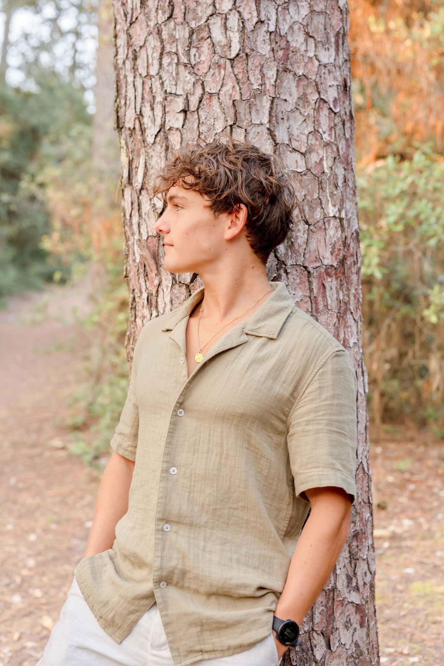 Person in casual beige shirt leaning against large pine tree trunk on forest path.
