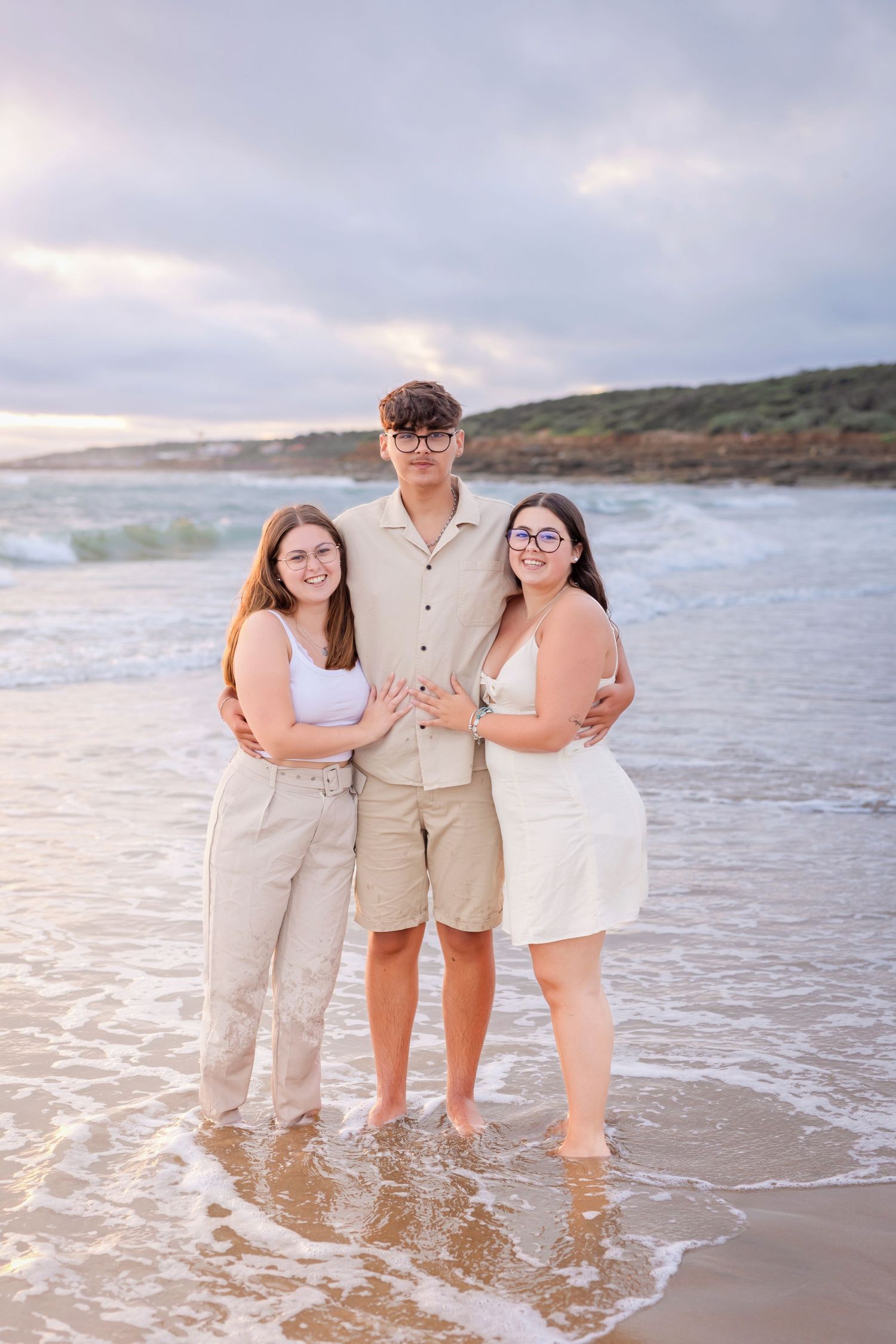 Three friends pose together on a beach with waves and cloudy sunset sky in the background.